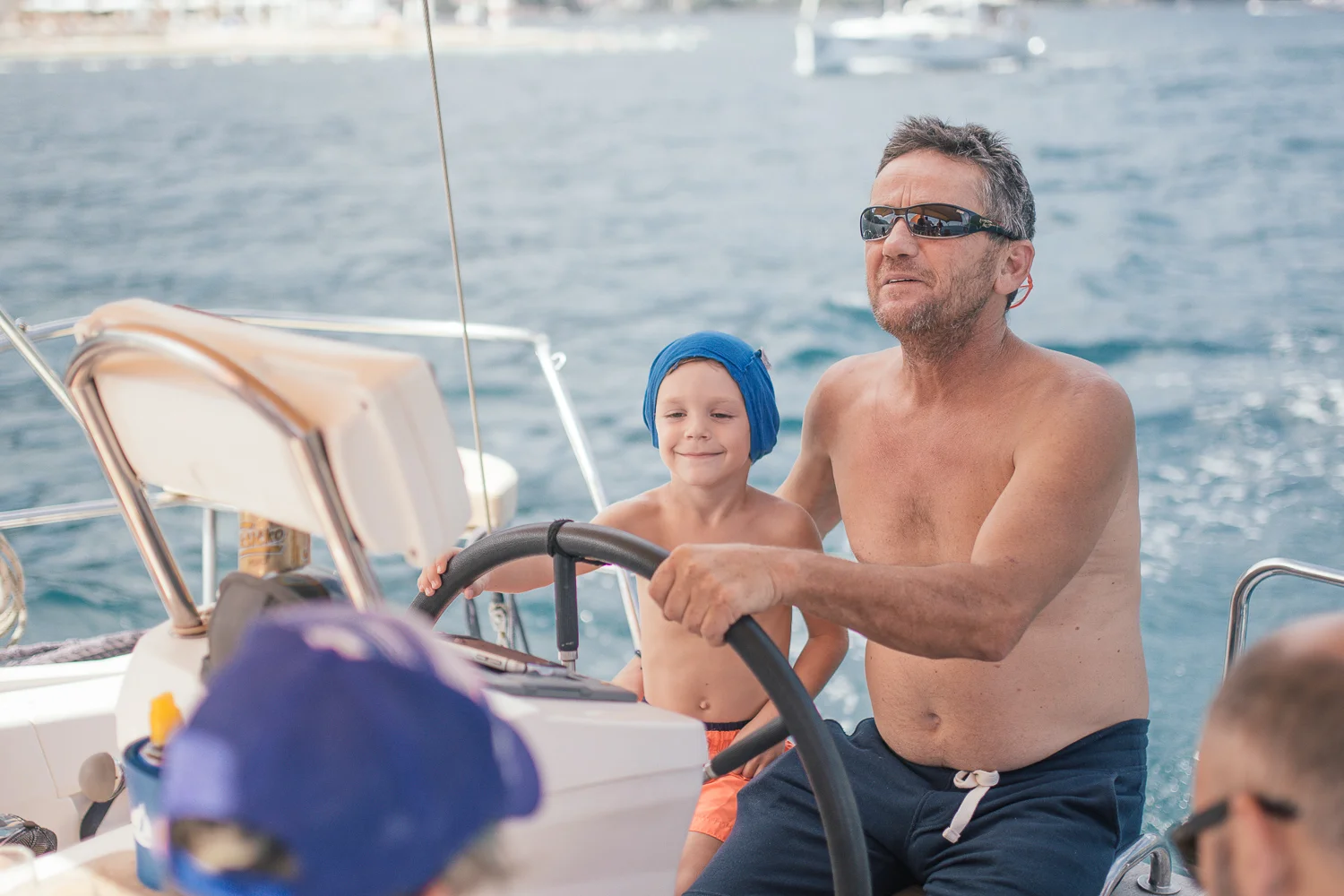 A man and a young boy on a boat, both shirtless, with the man wearing sunglasses and the boy wearing a blue head covering, steering the boat in the water.