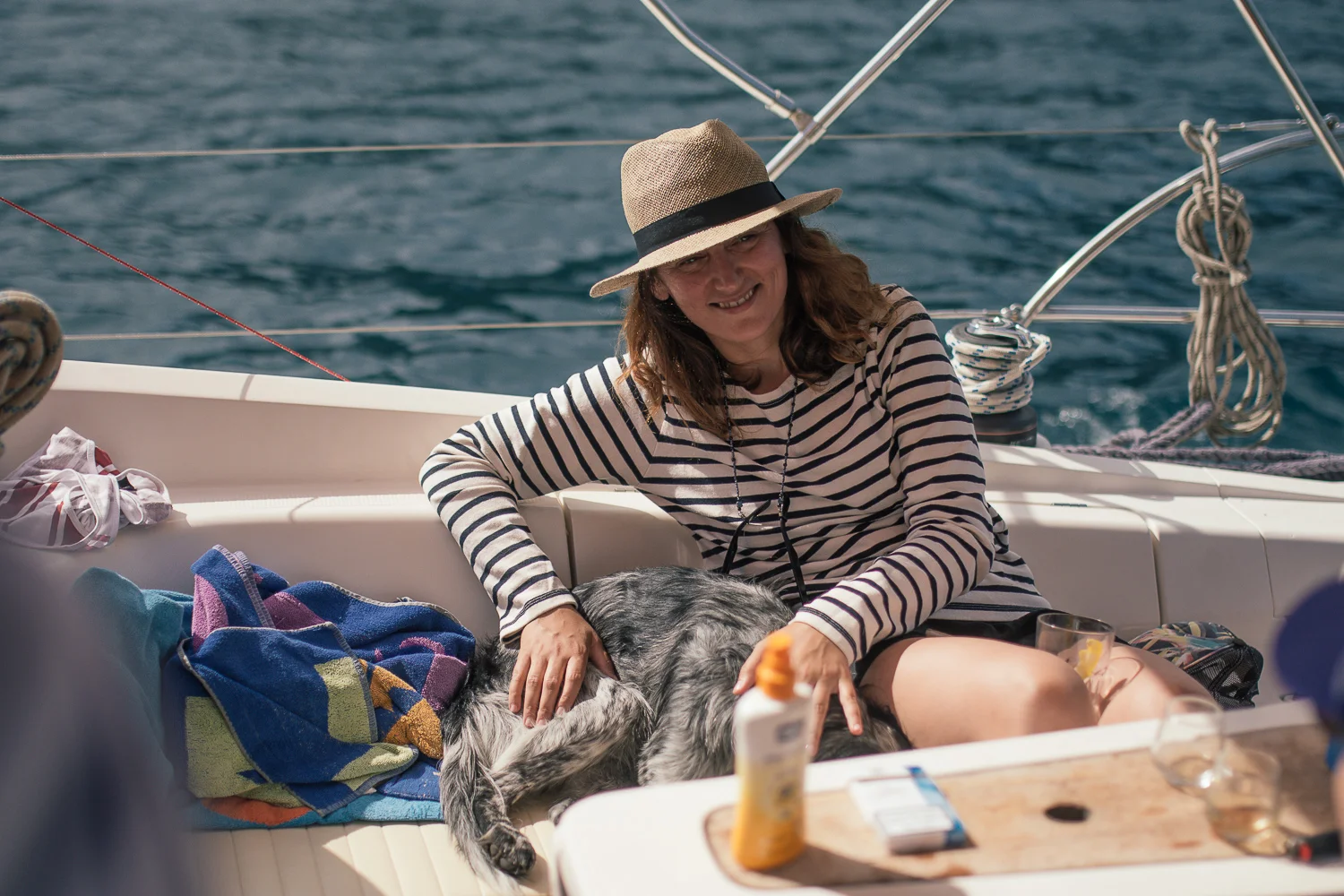A woman sitting on a boat with a dog, wearing a striped shirt and a straw hat, smiling as she pets the dog. The boat has water and sailing equipment visible in the background.