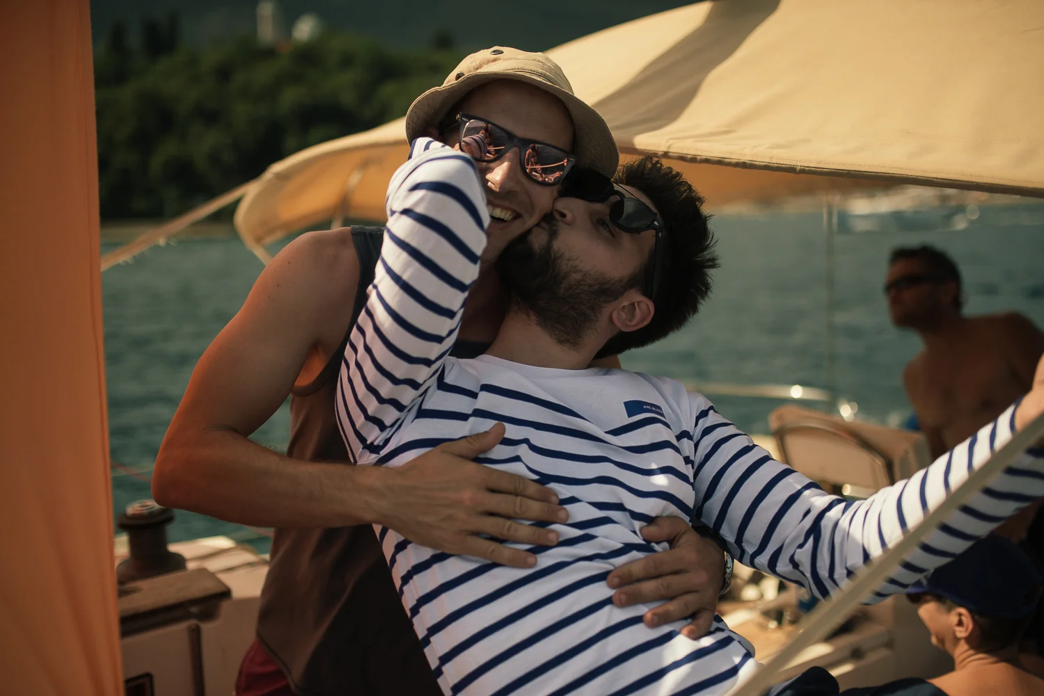 A couple enjoying a moment on a boat with the woman hugging the man from behind, both wearing sunglasses, with a man in the background on the boat and water and trees visible behind them.
