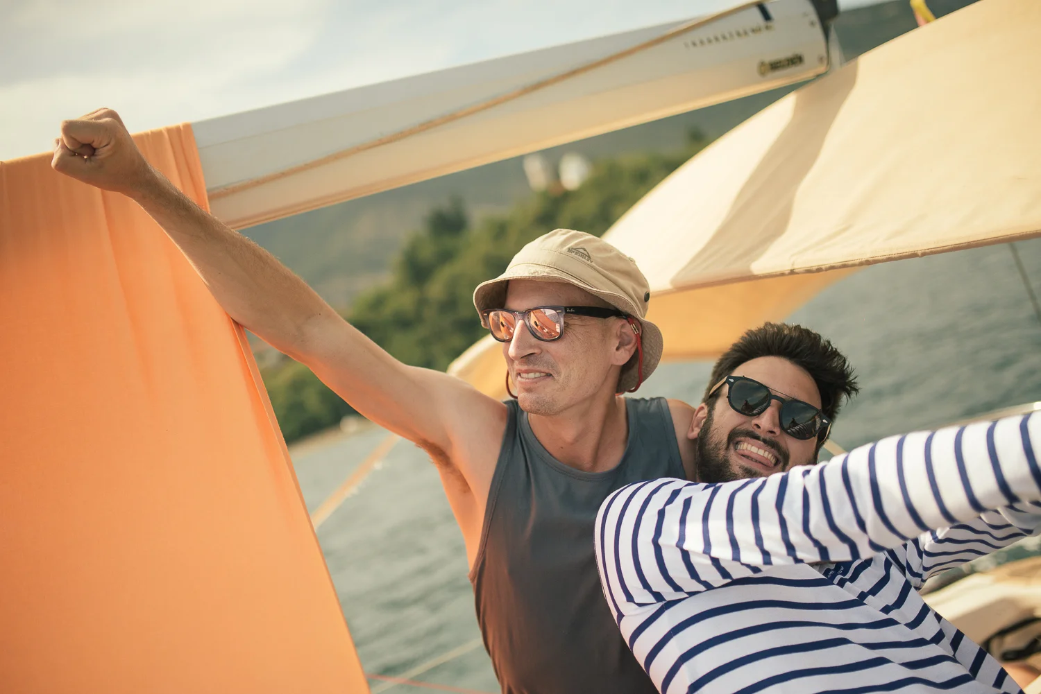 Two men on a boat taking a selfie, smiling, wearing sunglasses and casual summer clothes, with a lake and trees in the background.