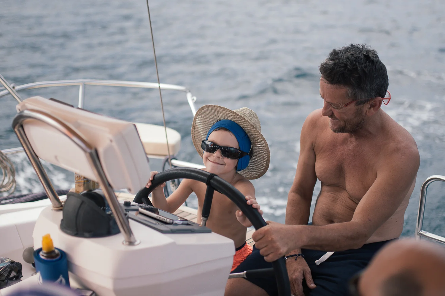A young boy and an older man are on a boat, smiling and enjoying the ride. The boy is wearing sunglasses, a sun hat, and headphones, while the man is shirtless with glasses, sitting beside him amidst the open water.