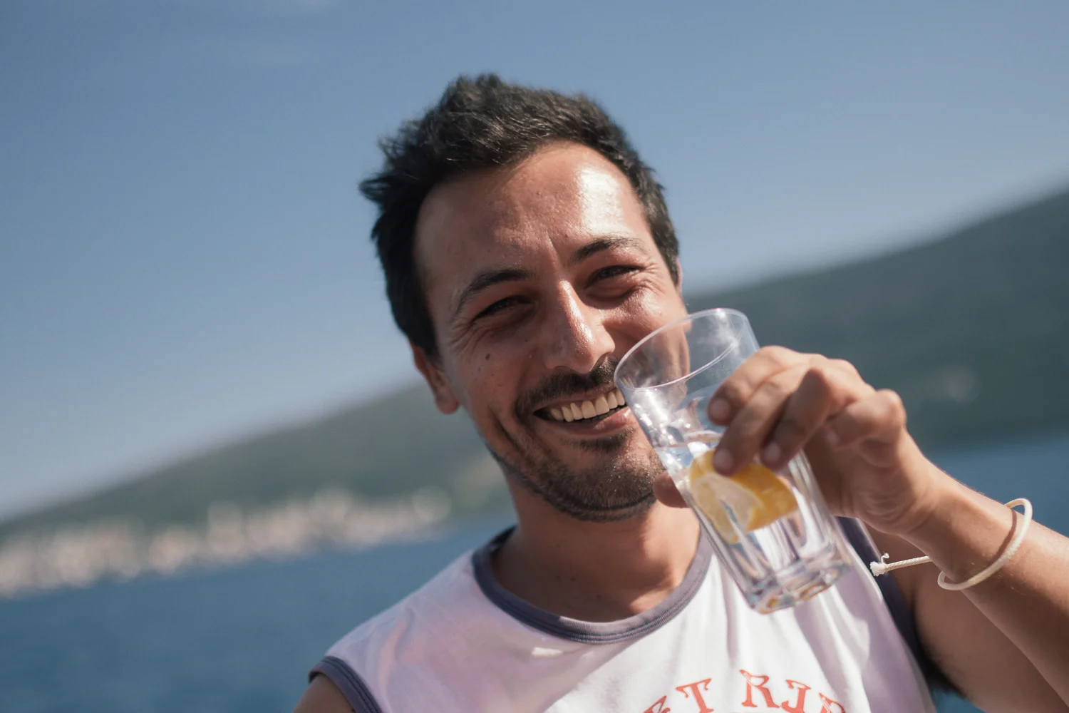 Smiling man holding a glass of water with lemon slices outdoors near water and hills in the background.
