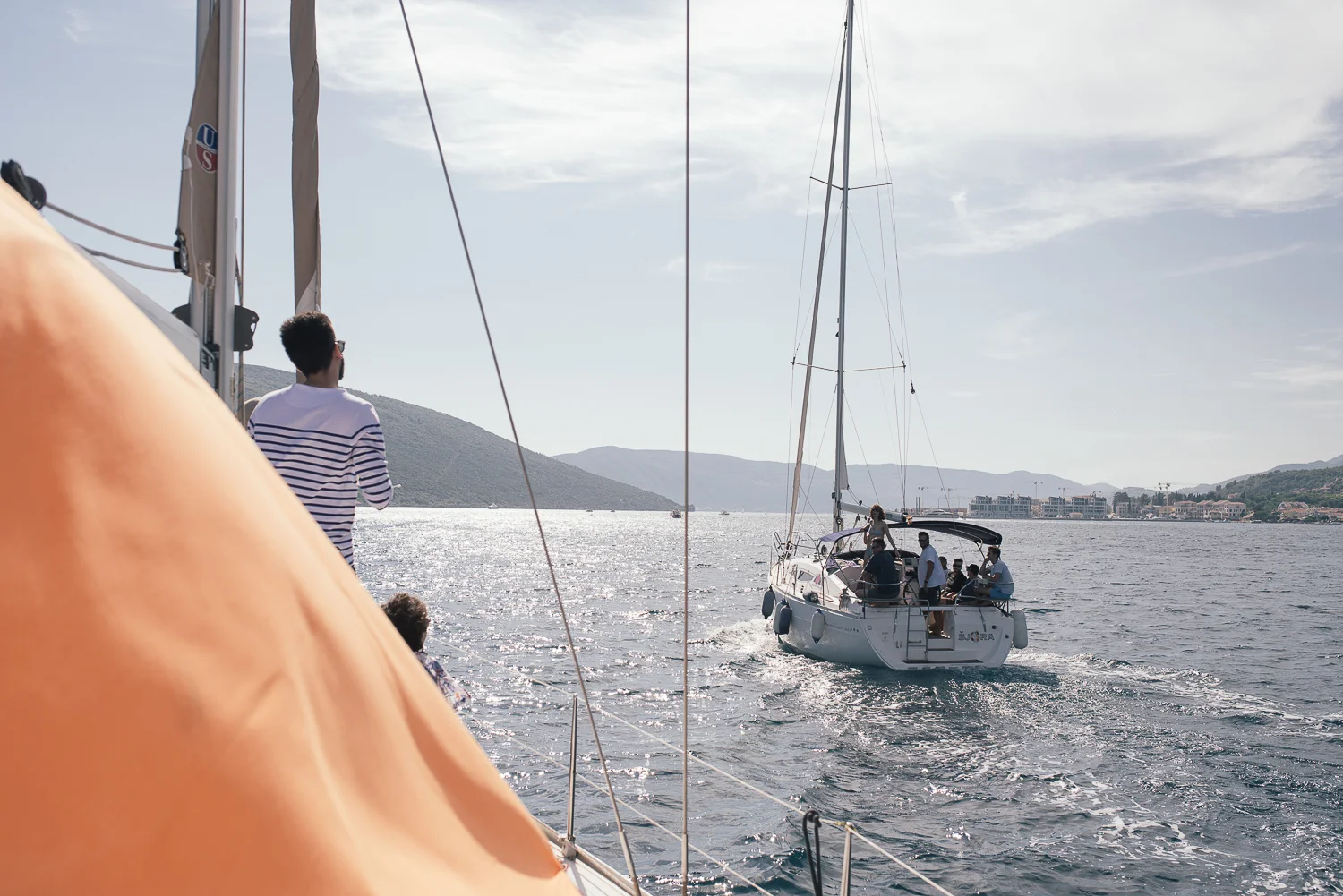 A group of people on a sailboat enjoying a day on the water, with some people looking towards another boat in the distance, on a sunny day with a scenic backdrop of hills and buildings.