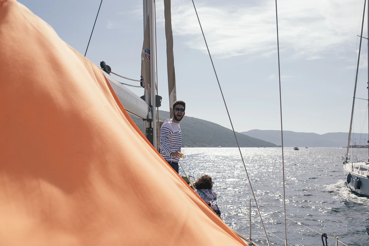 A man on a boat holding a drink, smiling, wearing sunglasses and a striped shirt, with water and mountains in the background on a sunny day.