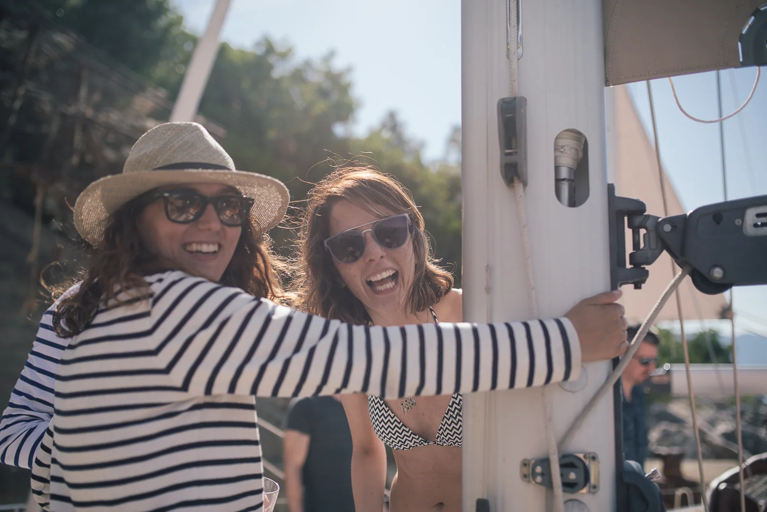 Two women in sunglasses and summer clothing smiling and having fun outdoors next to a boat with one woman turning a crank and the other laughing.