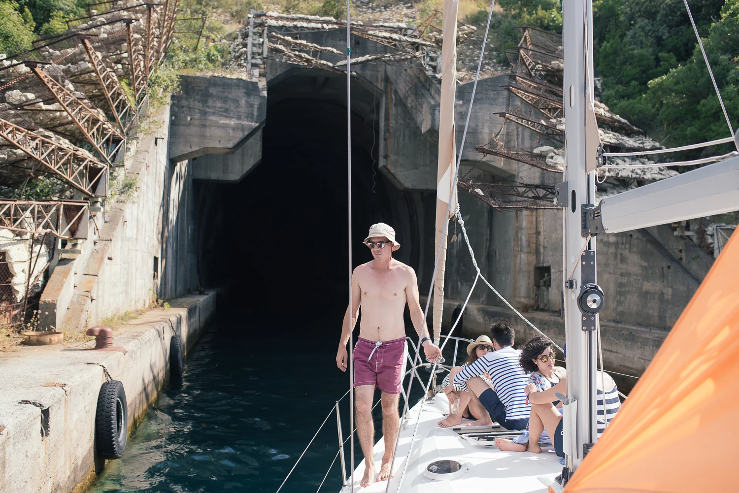 A group of people on a boat preparing to enter a dark tunnel at a waterway, with one man standing at the bow and others sitting or leaning on the boat's side.