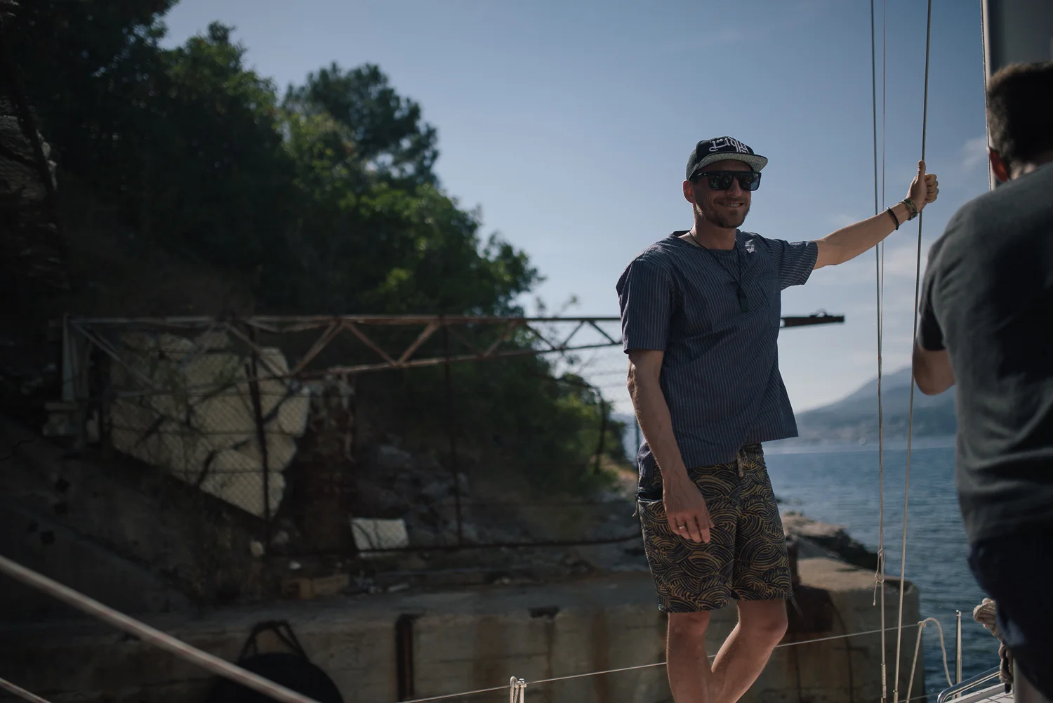 A man with a beard wearing sunglasses, a dark striped shirt, and patterned shorts standing on a boat, smiling, with another person partially visible holding a rope or pole. The background shows water, a rocky shoreline, and some trees.
