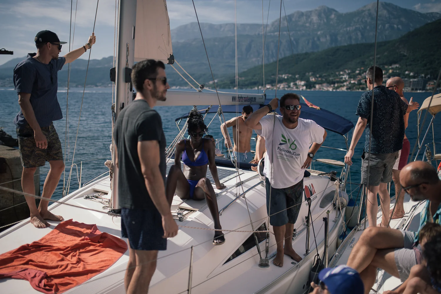 Group of people on a sailboat near a dock, with mountains in the background, some wearing casual attire, one woman in swimwear, and a man wearing sunglasses smiling and holding a rope.