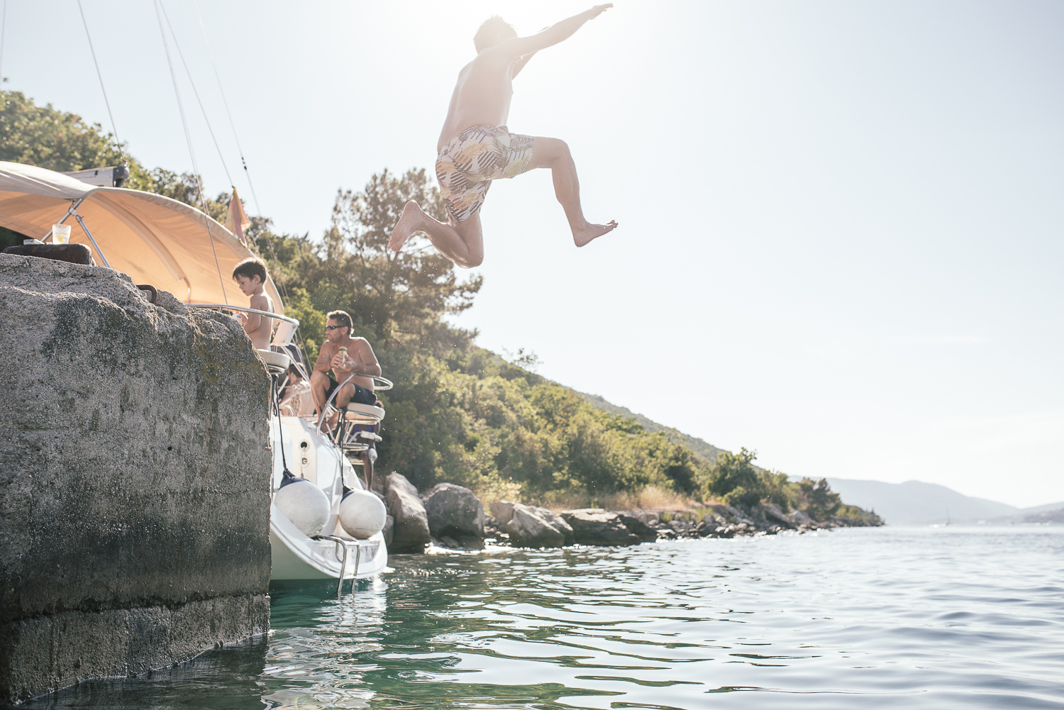 Person jumping off a boat into the water at a lake or seaside with people on the boat and trees in the background.