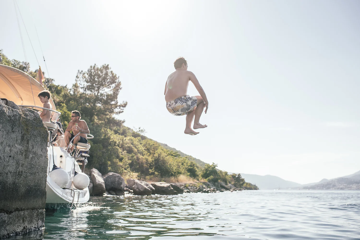 A man in patterned swim shorts jumps off a dock into a lake, with two boys and a man relaxing on the boat nearby, surrounded by trees and hills under daytime sun.