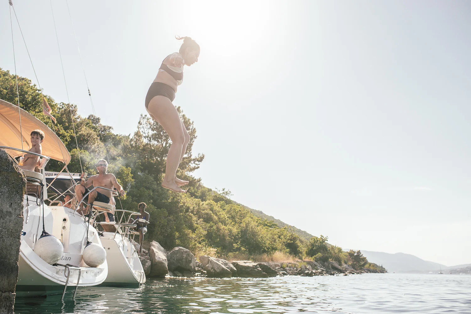 A woman jumping off a boat into the water, with three people on the boat watching, on a sunny day near a wooded shoreline.