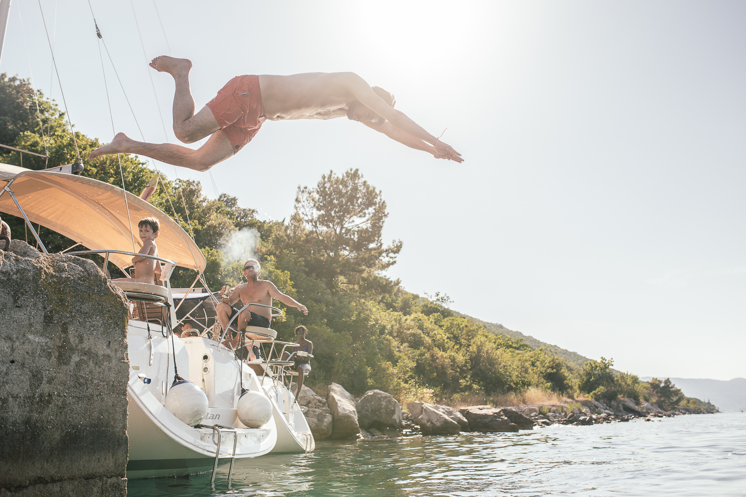 A man diving off a sailboat into the water near a rocky shoreline with trees, with other people on the boat and a sunny sky.