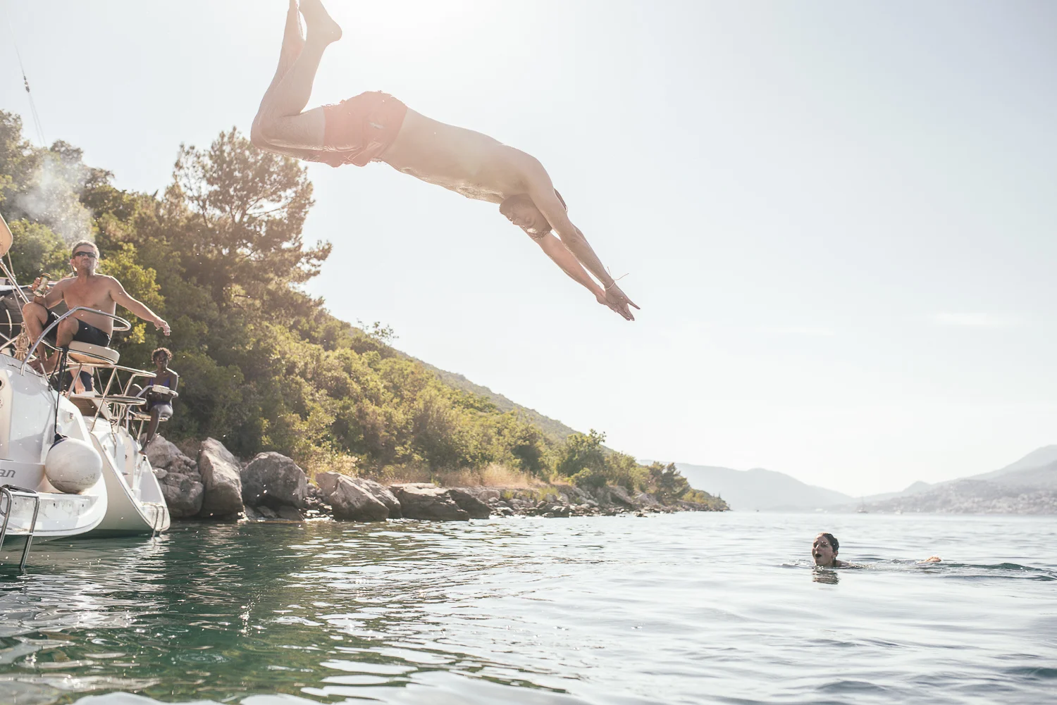 A man jumping off a boat into a lake while others watch from the boat and swim in the water, with trees and mountains in the background during daytime.