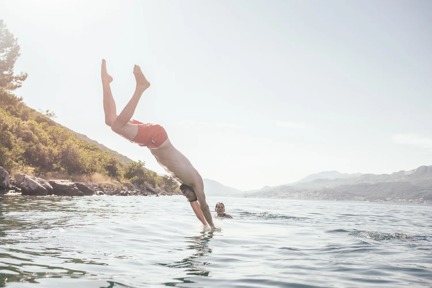 A man performing a handstand dive into a body of water with a woman swimming nearby, surrounded by mountains and trees in the background.