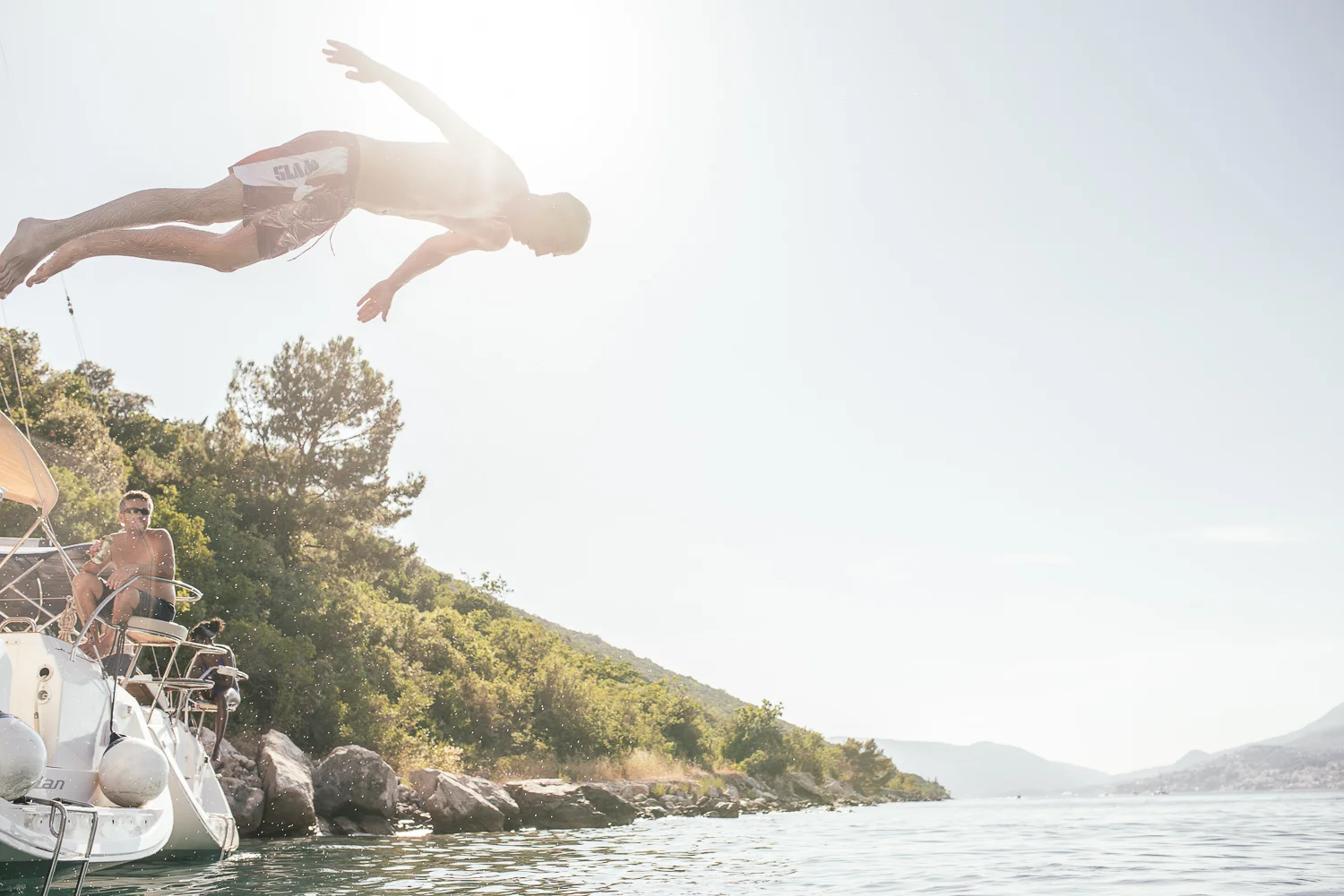A person is diving off a boat into a body of water with another person sitting on the boat, surrounded by trees and hills in the background in a sunny outdoor setting.