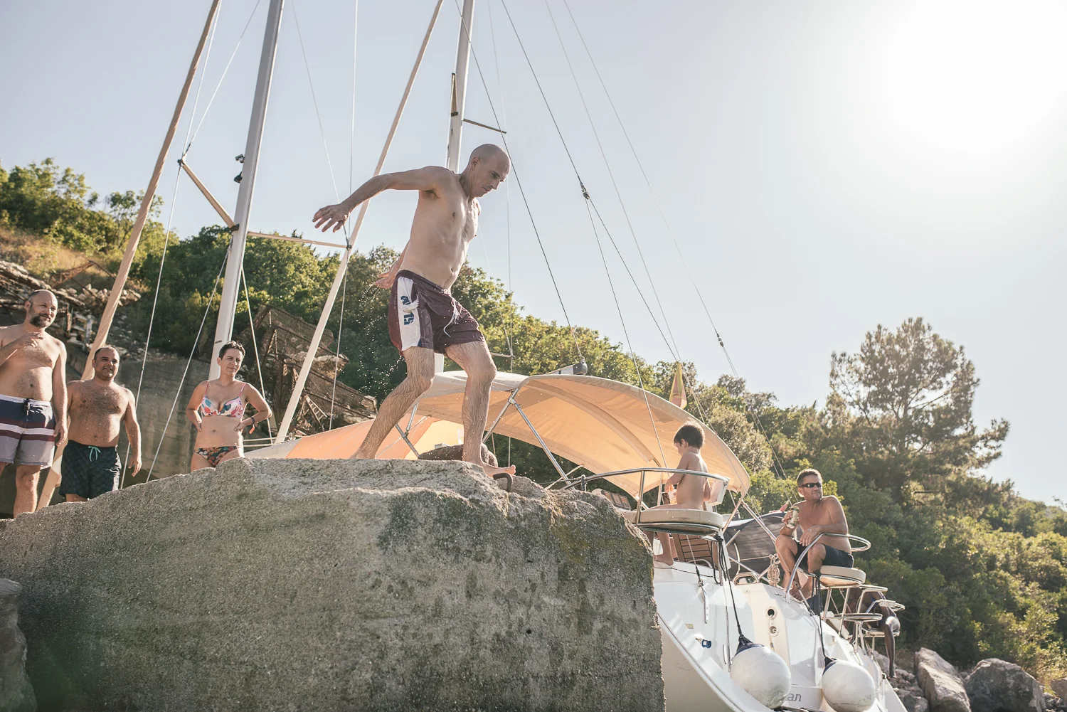 A group of people on a boat at a rocky shoreline with lush green trees in the background, and one man jumping into the water.