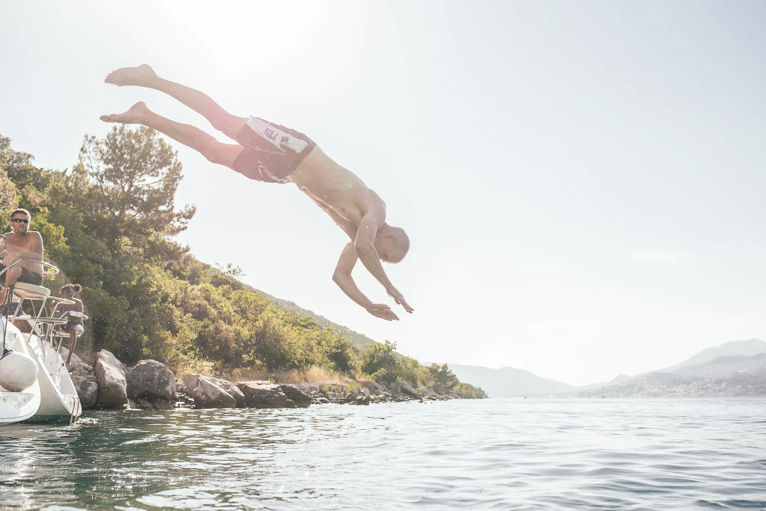 A man diving into the water from a boat, with a few other people on the boat, on a sunny day near a rocky and green lakeshore.