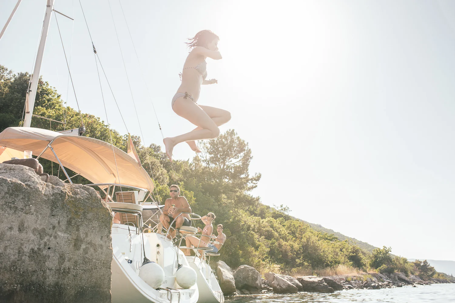 A woman jumping off a sailboat into the water with three other people on the boat enjoying a sunny day by the water surrounded by trees and rocks.