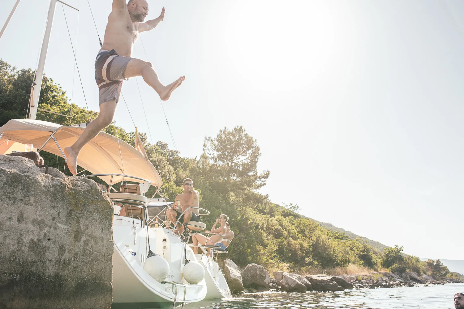 A man jumping off a boat into the water while three people sit on the boat by the shore, with trees and rocks in the background during bright, sunny weather.