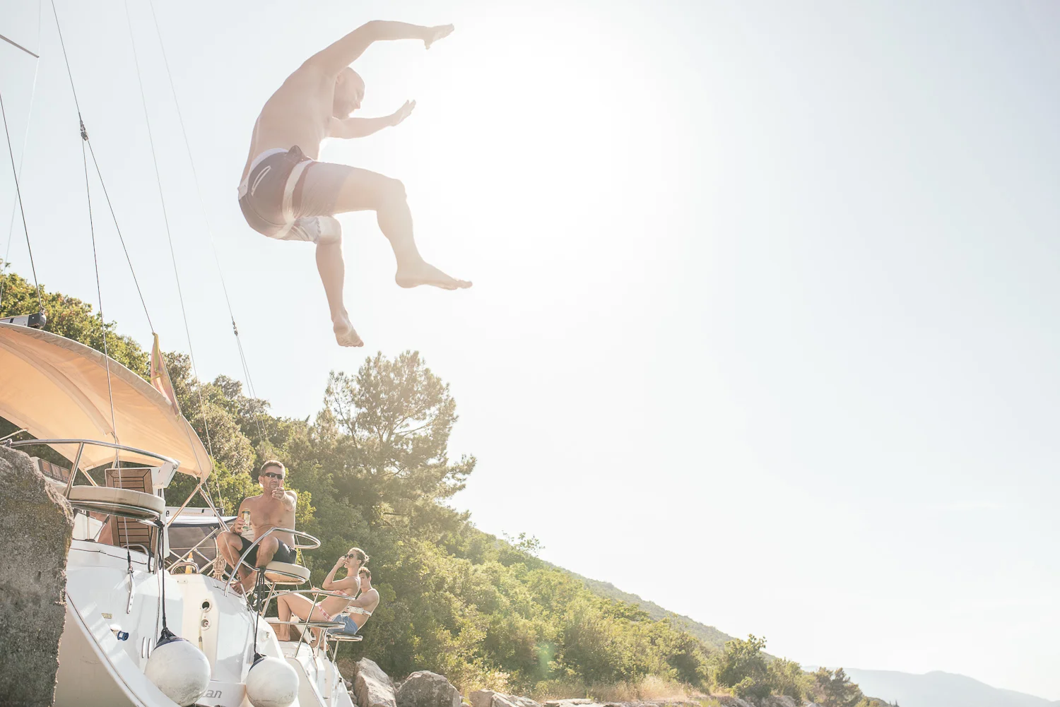 A person jumping off a boat into the water while other people are relaxing on the boat in a scenic outdoor setting.