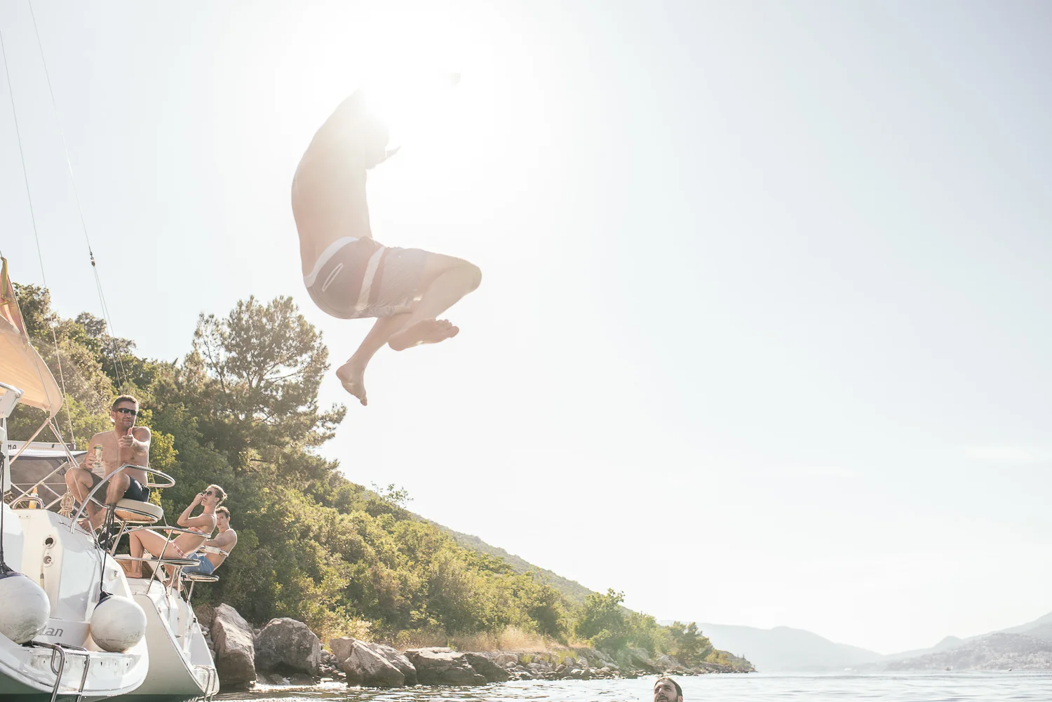 Person in mid-air diving from boat into a river with lush greenery and hilly landscape in background, and a group of people on the boat watching and taking photos.