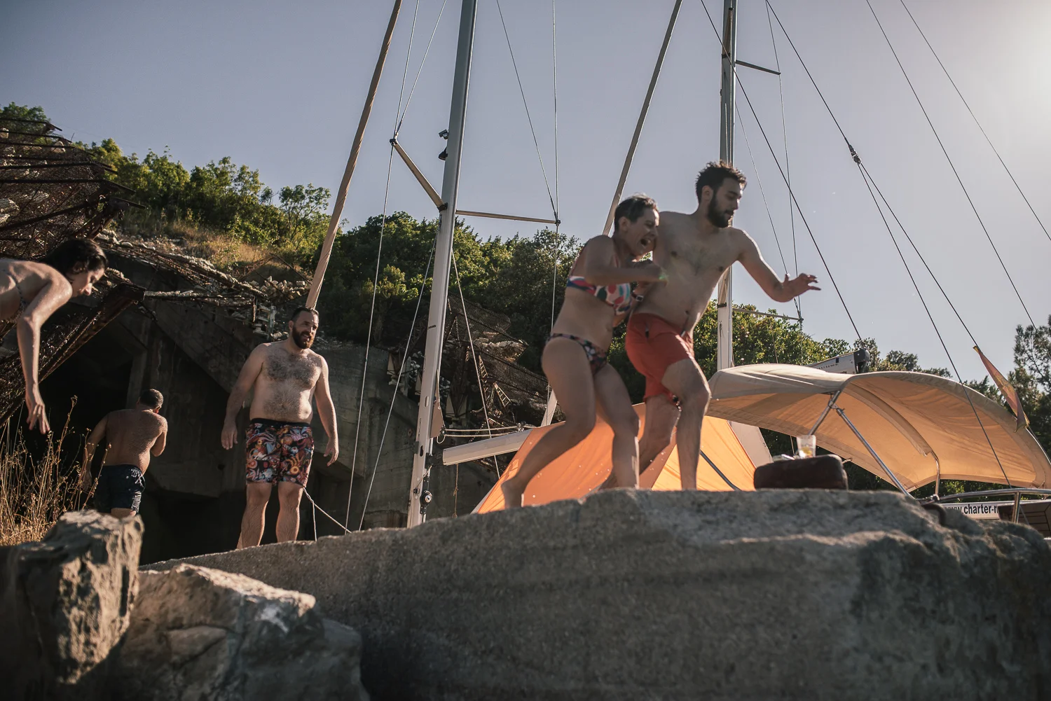 Group of adults enjoying a swim and fun at a lakeside or beach area with boats and rocky shoreline, during sunny weather.