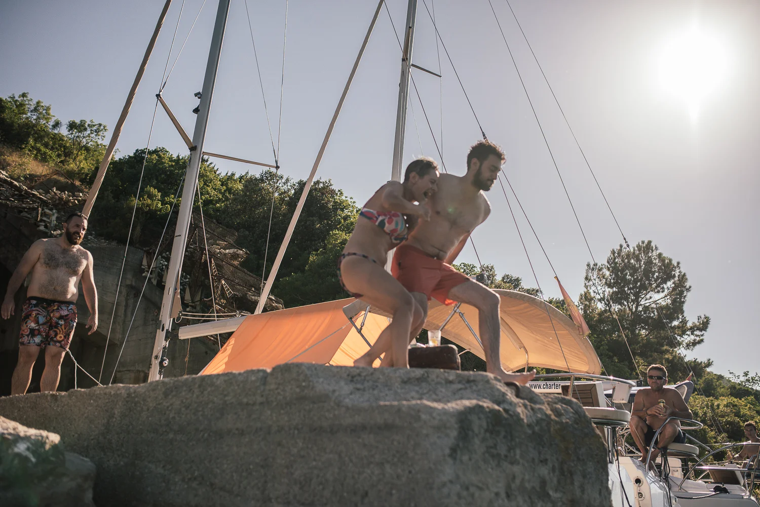 Group of friends enjoying themselves on a boat and rocky shore during a sunny day, with some jumping off the rock into the water, others sitting on the boat, and a man walking along the shore.