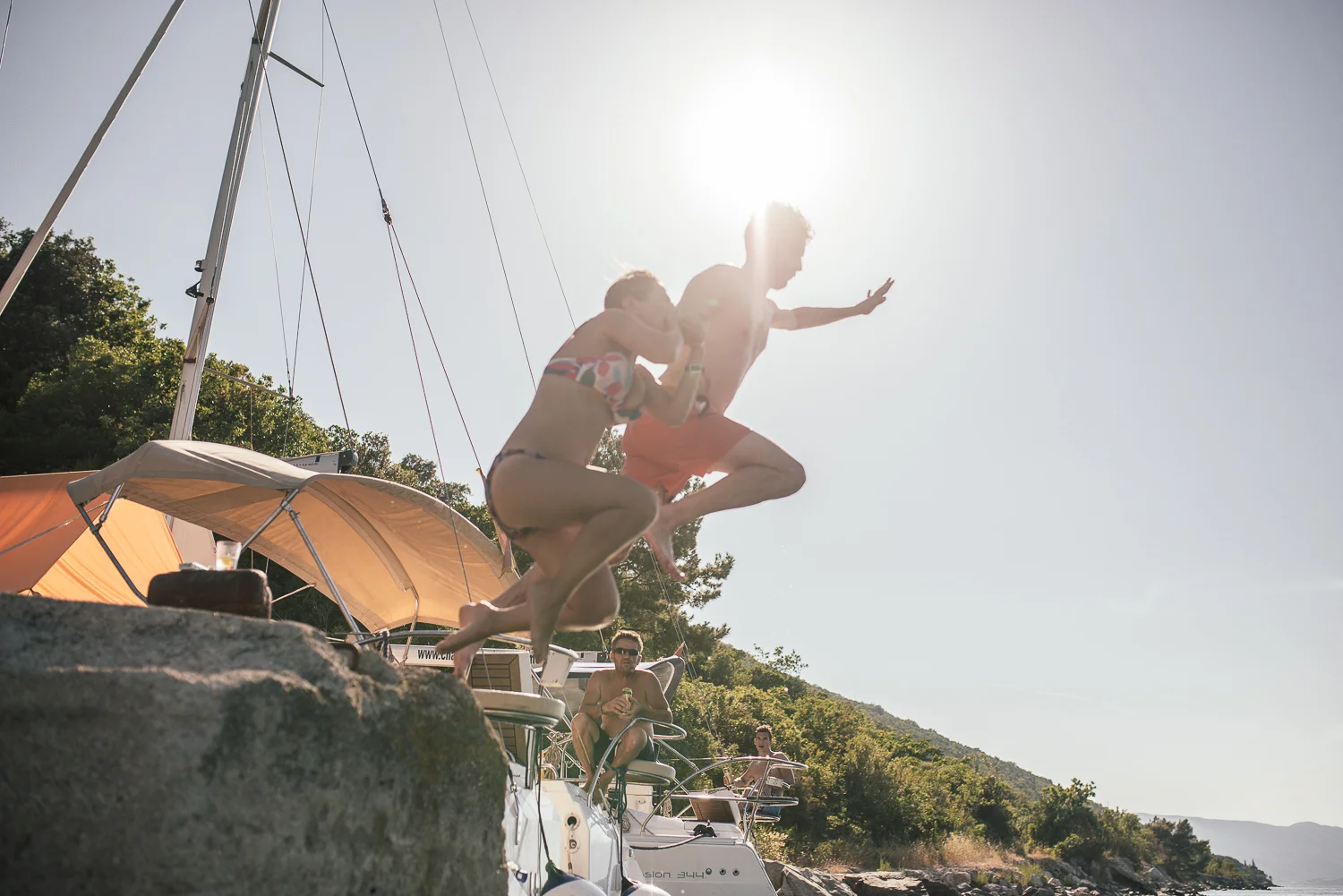 Two young men in swimsuits jumping off a boat into the water with other people on the boat, boat umbrellas, and trees in the background on a sunny day.