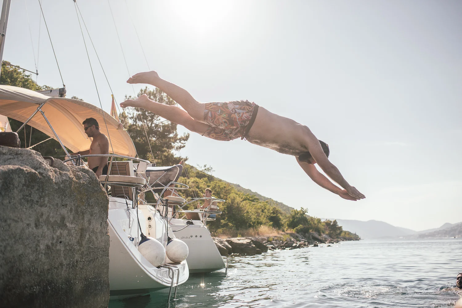 A man jumping into the water from a boat near a rocky shoreline during daytime with other people on boats and trees in the background.