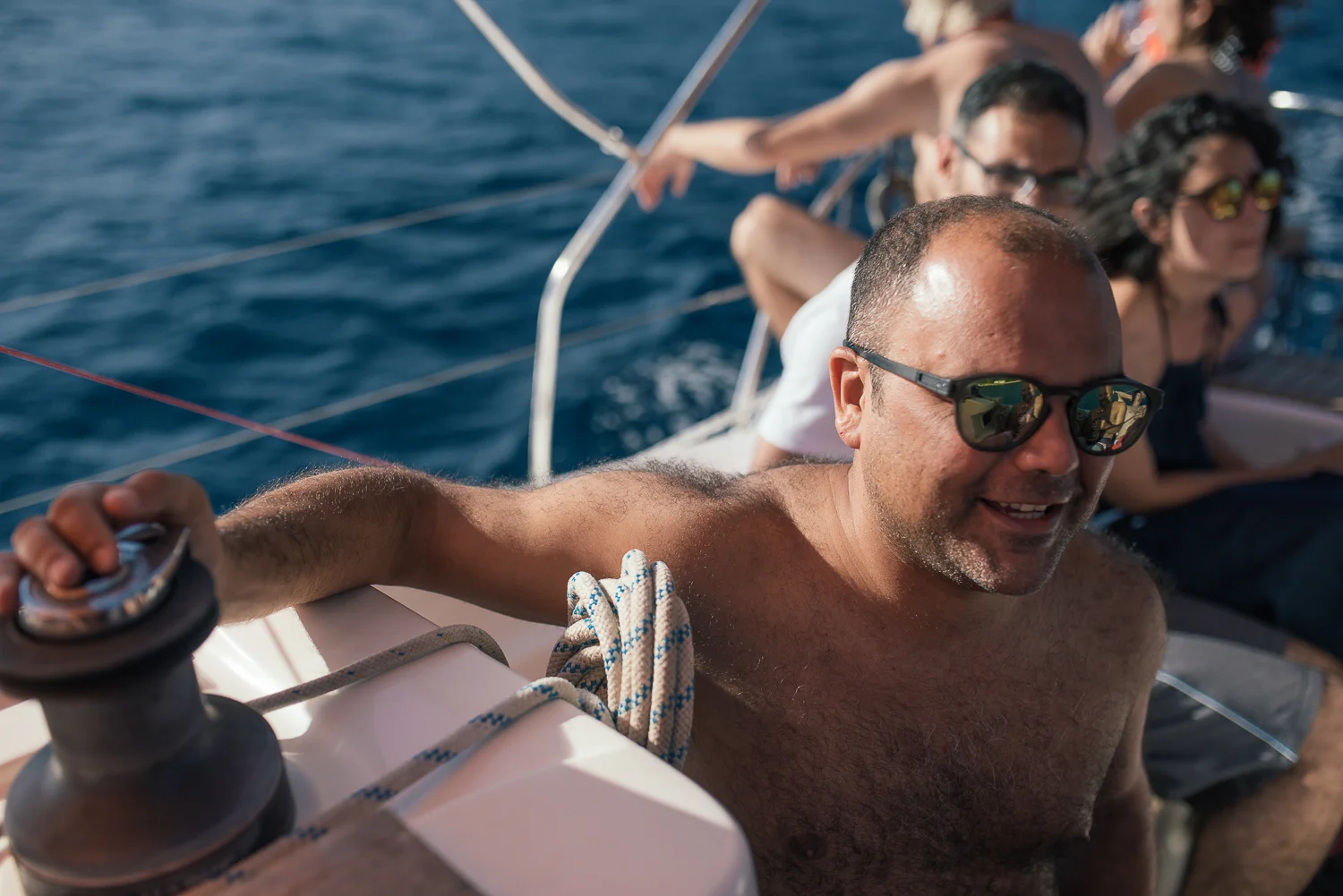 Man with sunglasses steering a sailboat, with other people sitting behind him, all enjoying a day on the water.
