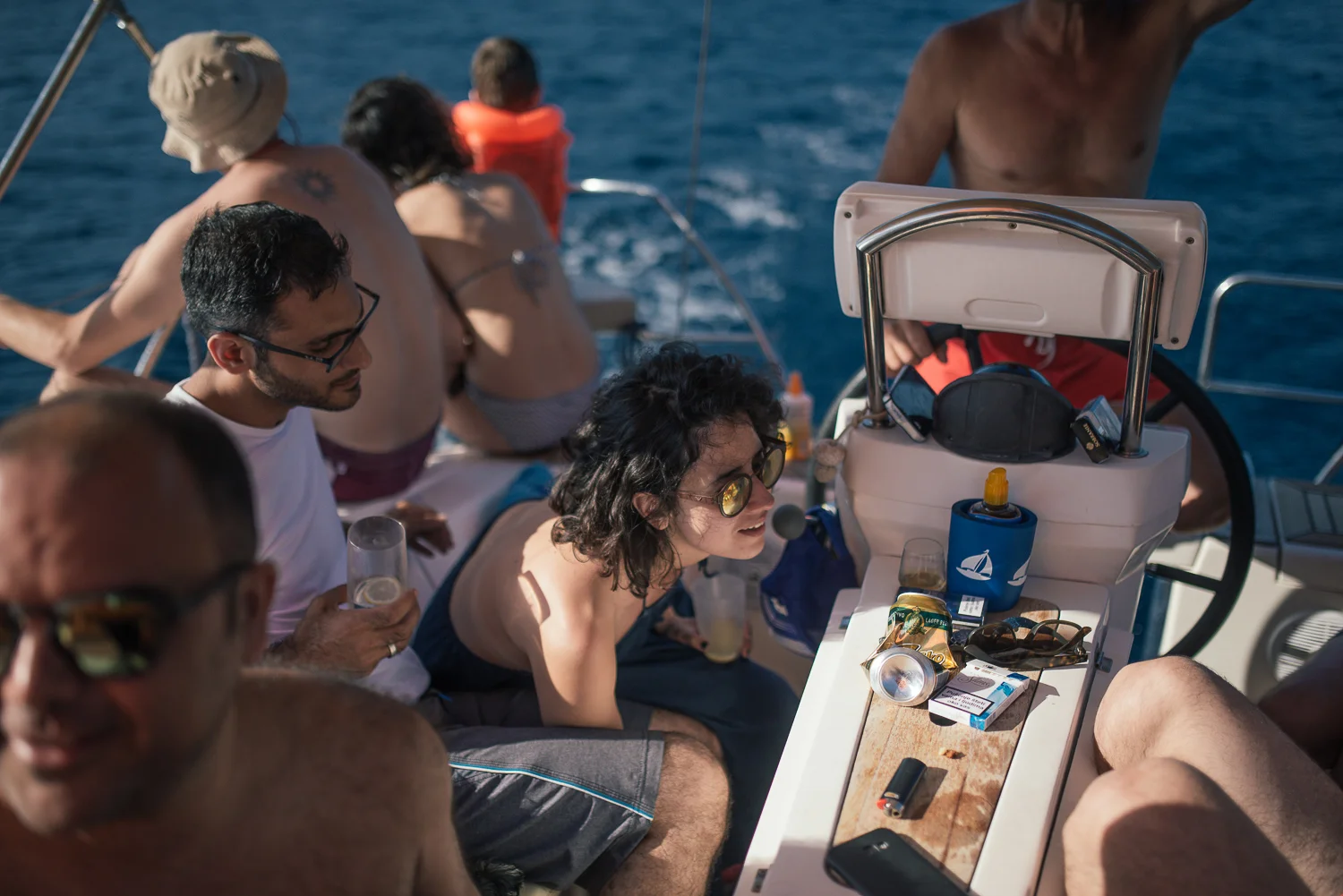 Group of people on a boat enjoying a sunny day, some are shirtless and wearing sunglasses, with drinks and snacks on a table near the helm.