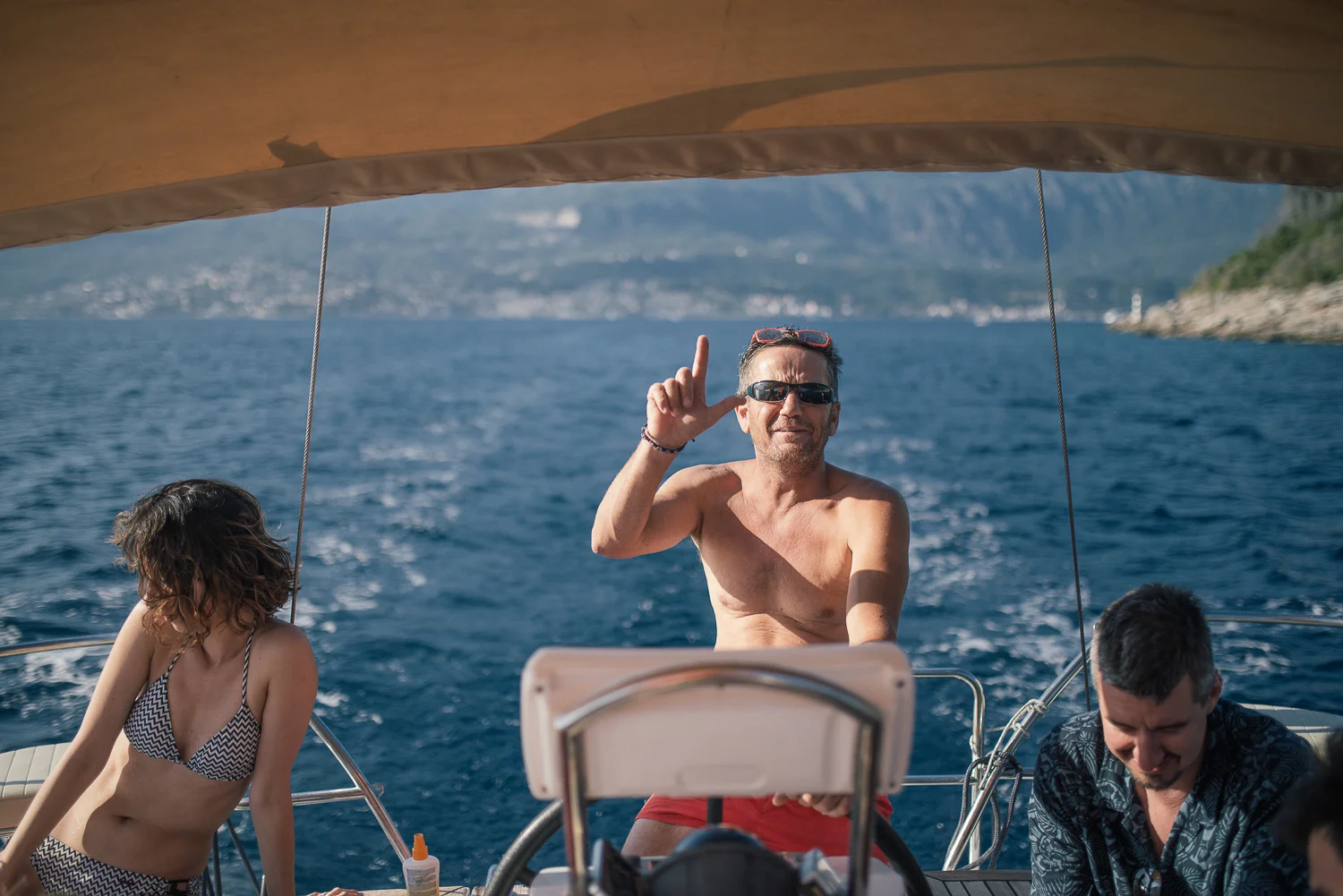Three people on a boat in the ocean with mountain scenery in the background. The man at center is shirtless, wearing sunglasses, and making an 'L' shape with his hand, smiling. A woman in a bikini and a man in a patterned shirt are on either side, lo