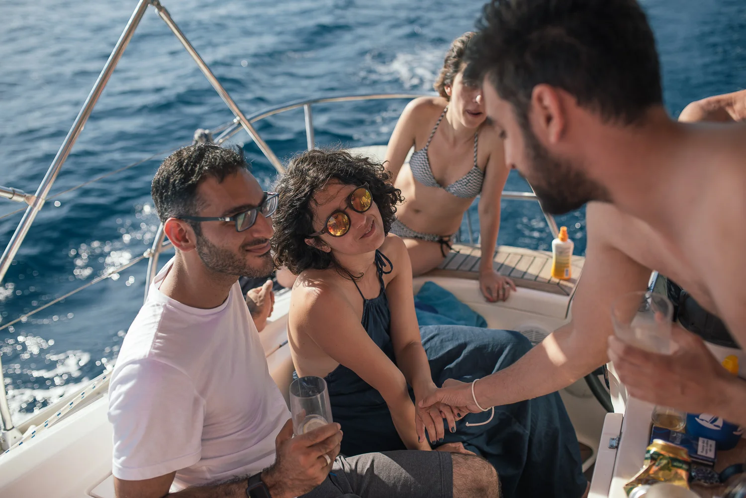 Group of friends on a boat at sea, engaged in conversation, with drinks in hand, during daytime with clear weather.