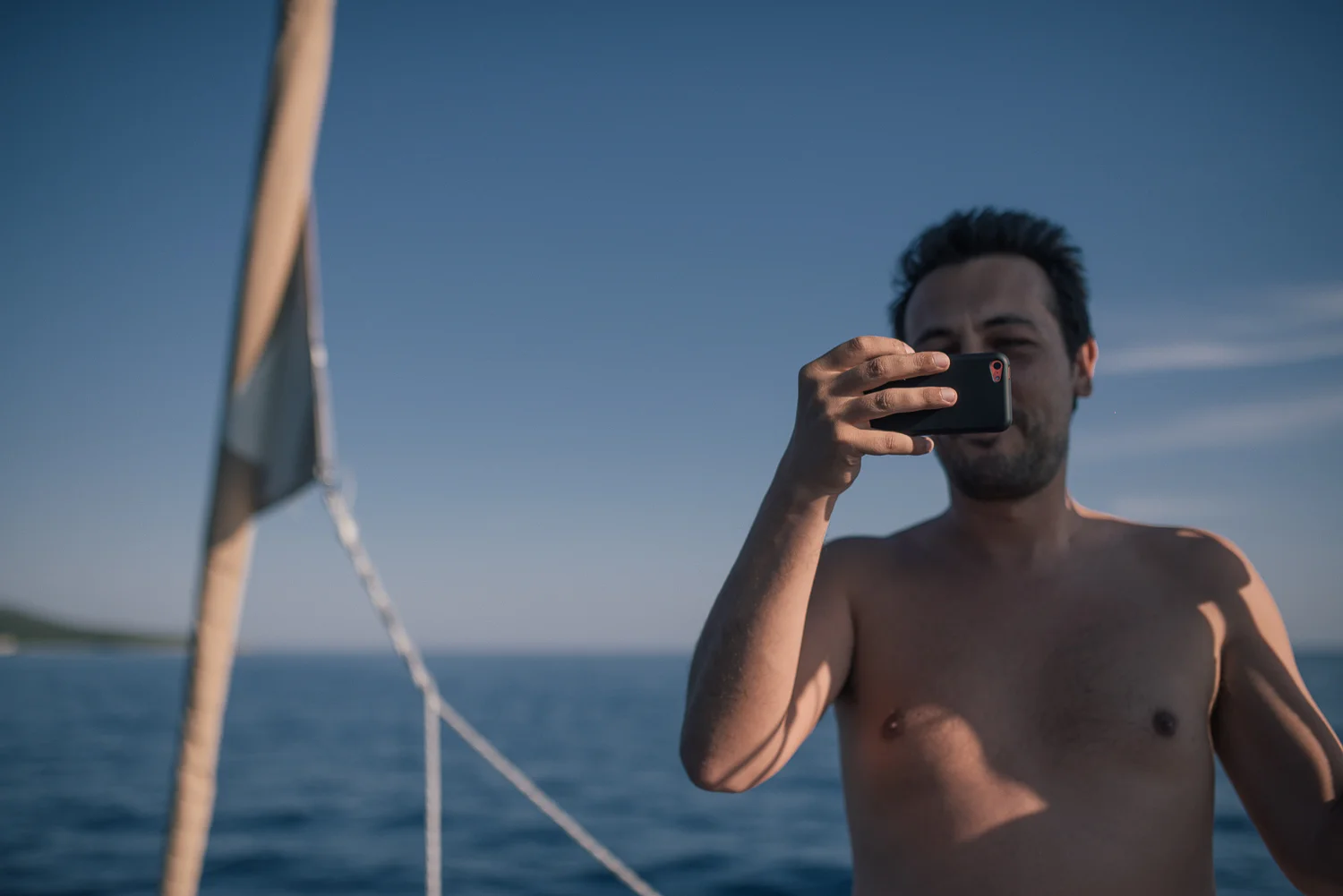 A shirtless man with dark hair taking a selfie on a boat with a smartphone, with an ocean and clear sky in the background.