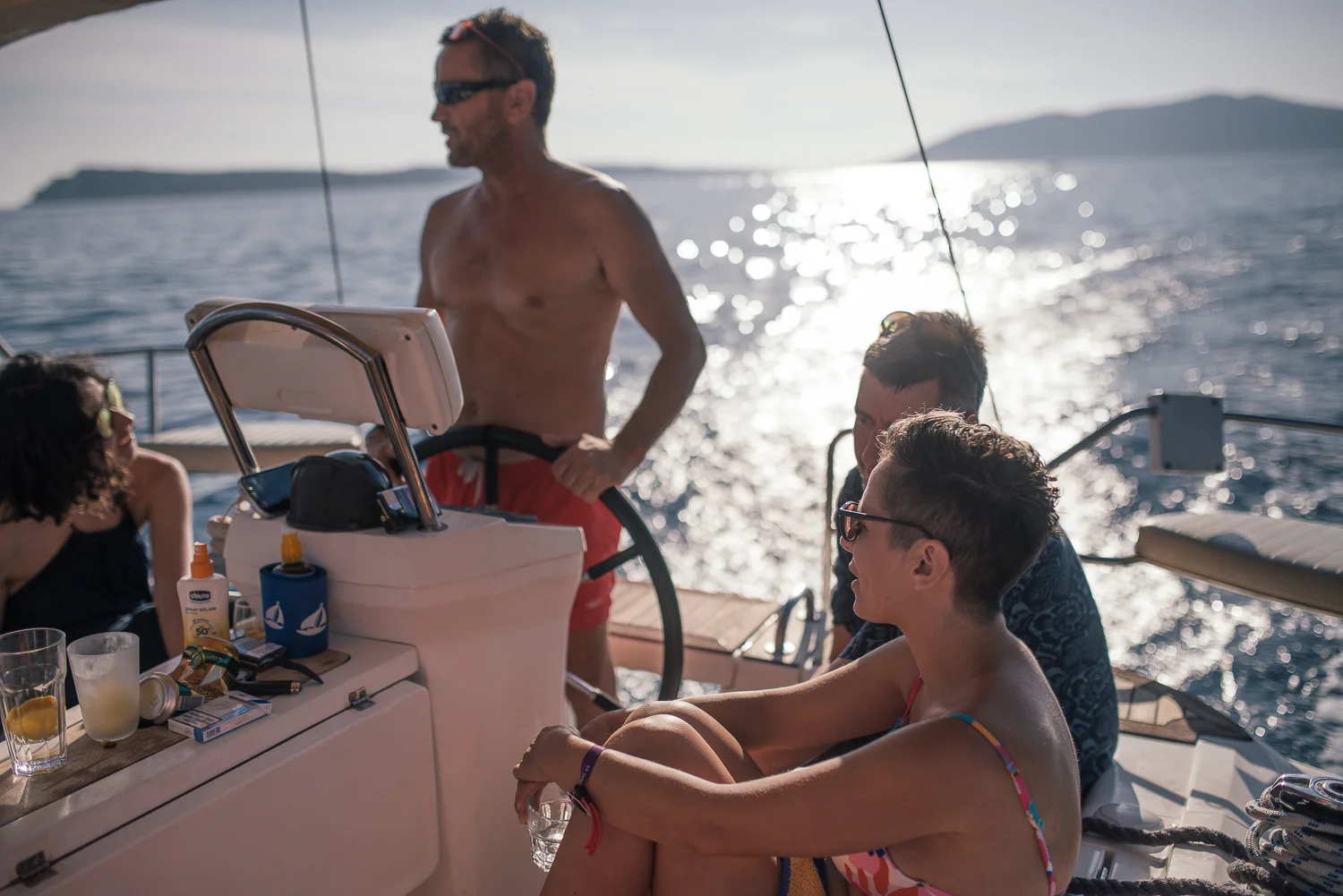 Group of friends enjoying on a boat during sunset, with water and an island visible in the background, some sitting and one standing at the steering wheel.