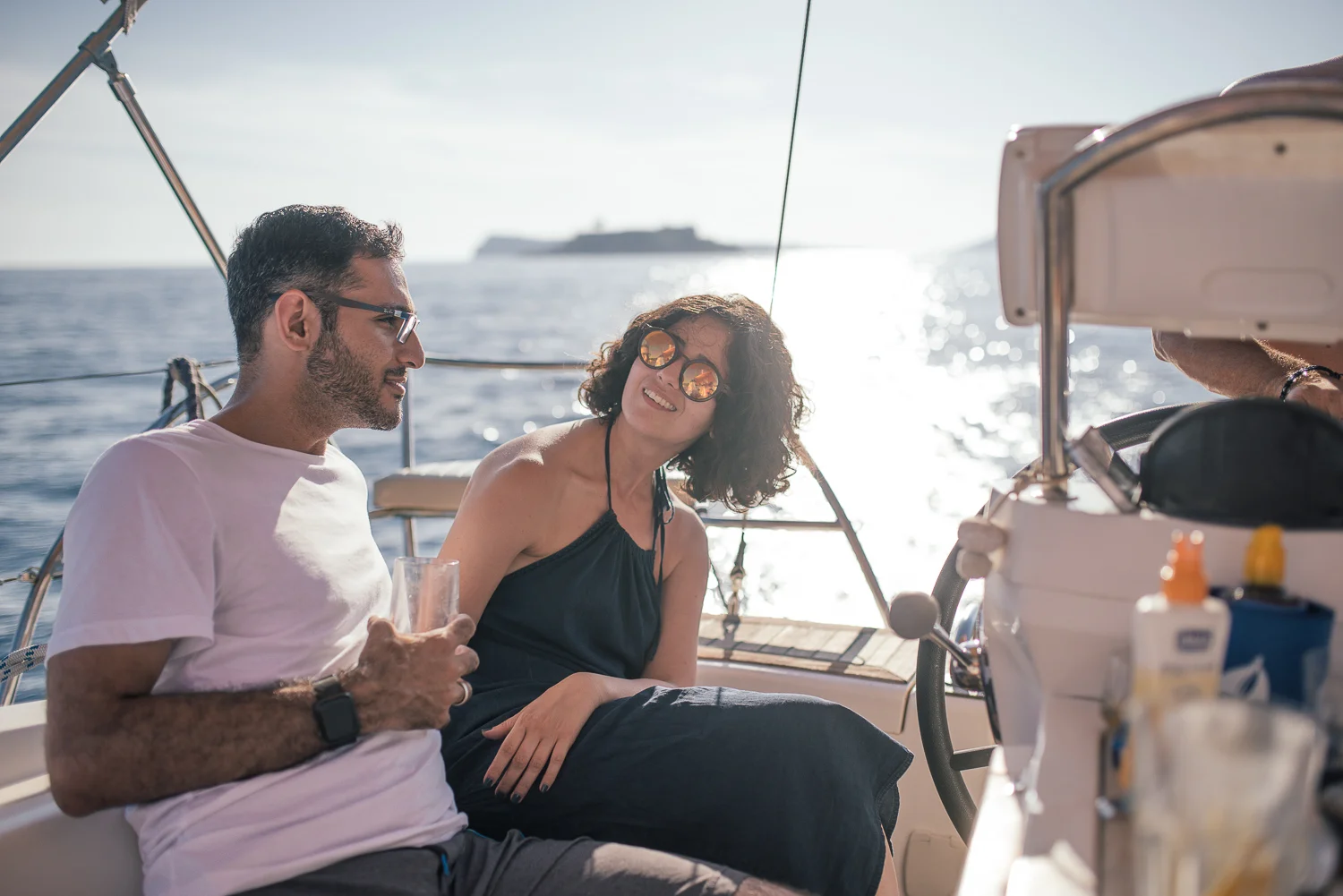 Two people on a sailboat enjoying sunny weather, with the man holding a glass and the woman smiling, while the ocean and a distant ship are visible in the background.