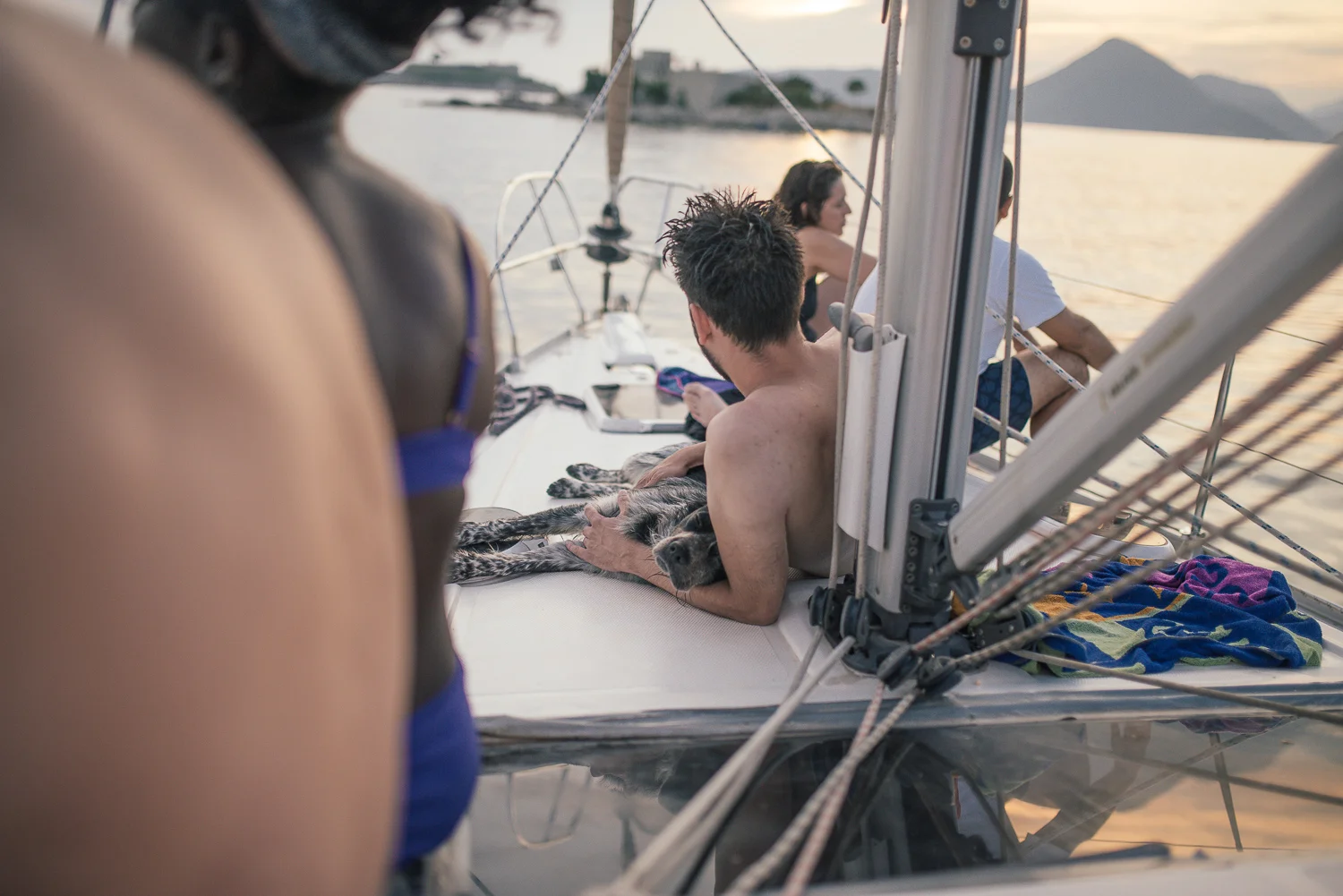 People relaxing on a sailboat at sunset, with a man petting a dog and two other individuals sitting and gazing at the water.