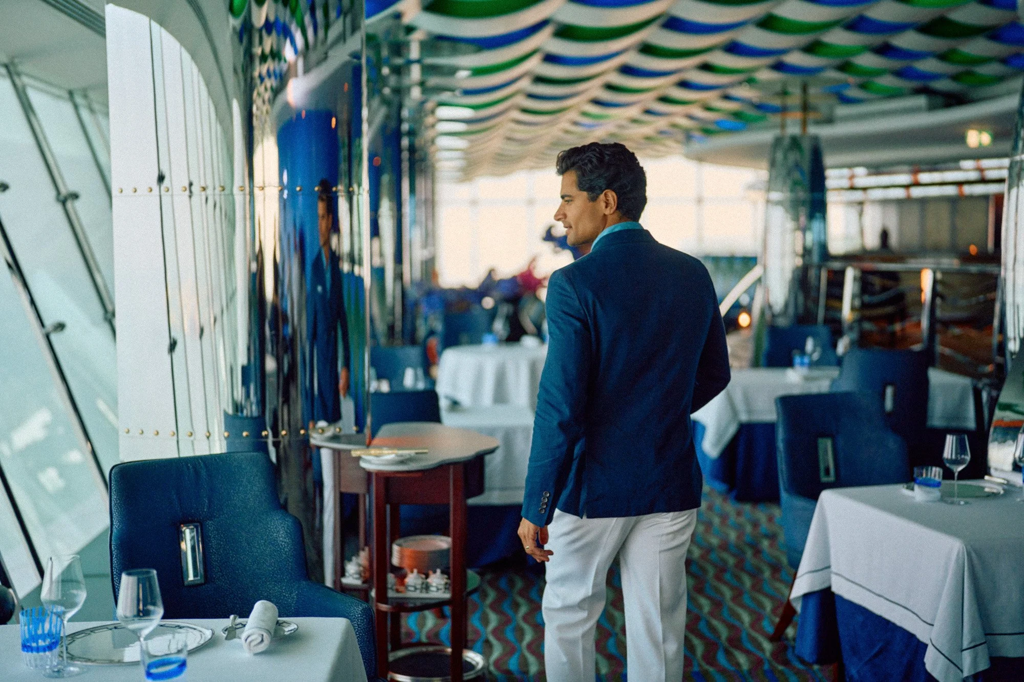 A man in a blue blazer and white pants inside a restaurant or dining area with tables set for a meal, some glasses and rolled napkins on the tables, and large windows with a colorful ceiling decoration.
