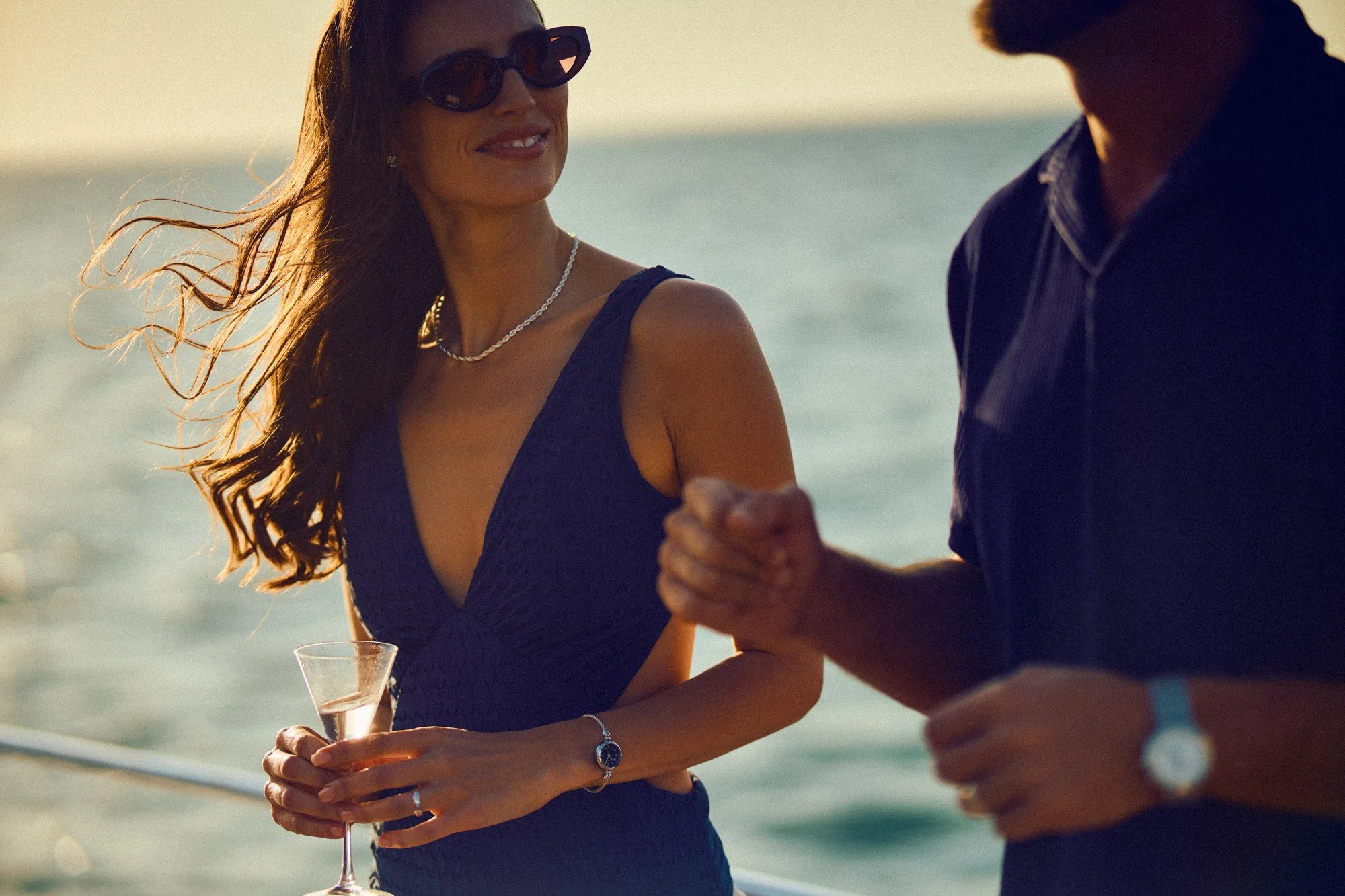 A woman in a dark blue dress and sunglasses holding a glass of wine, smiling and looking at a man, with a beach and ocean in the background during sunset.
