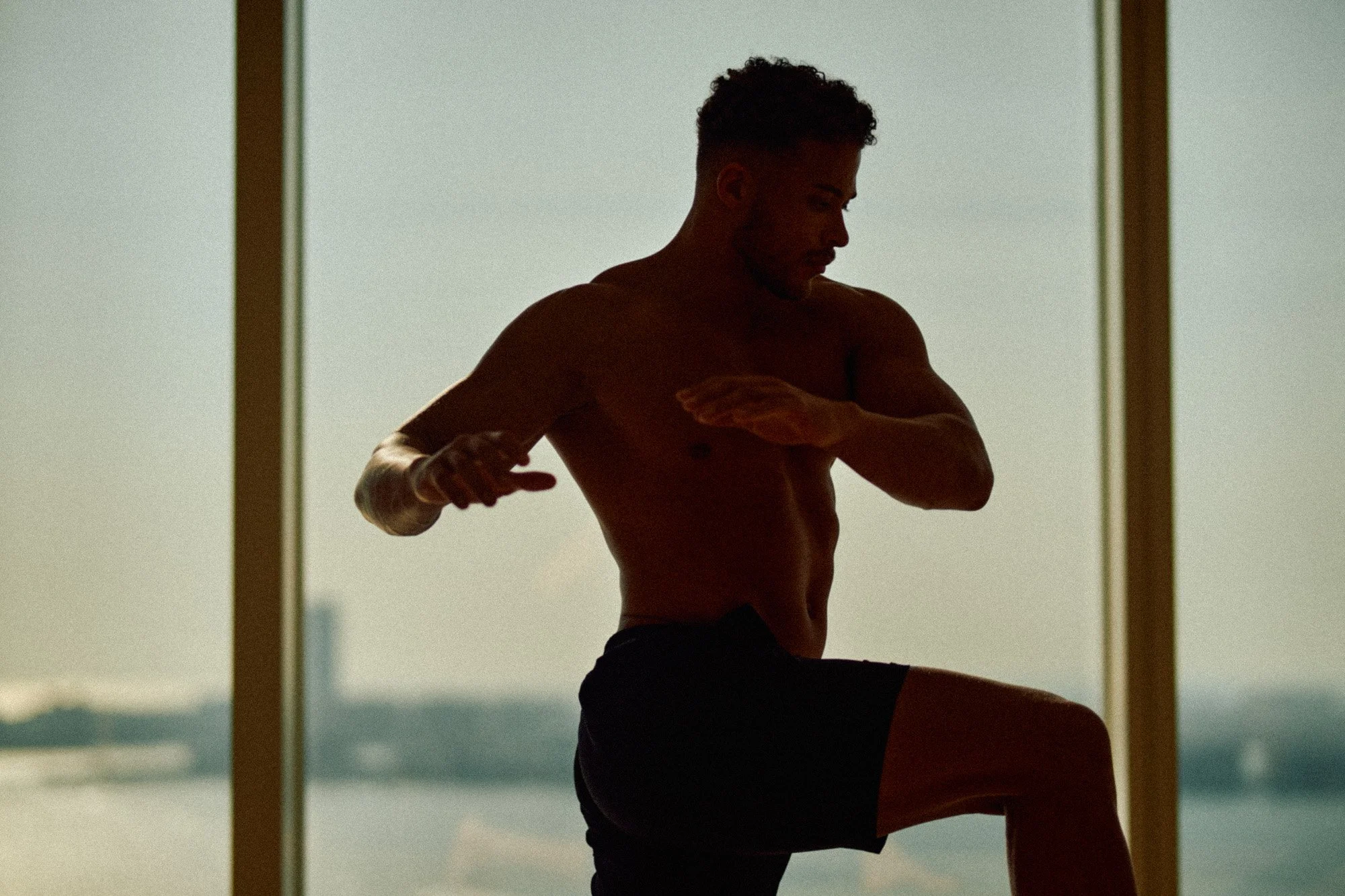 A shirtless man with a tattoo on his right arm is in profile, stretching or exercising in front of large windows with a view of water and a cityscape in the background.