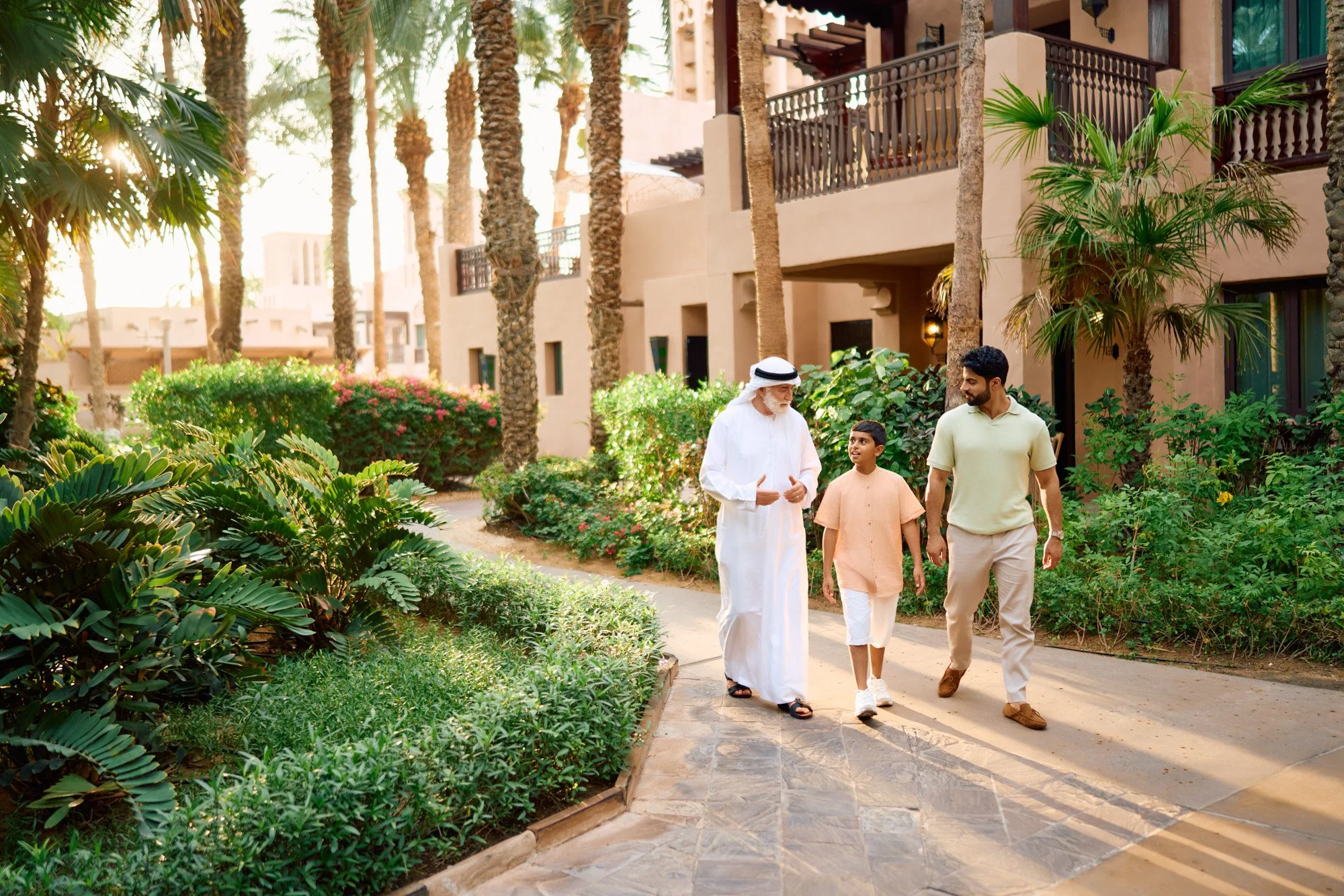 Three men walking and talking along a paved path in a lush, tropical garden area with palm trees and greenery, in front of a beige building with balconies.