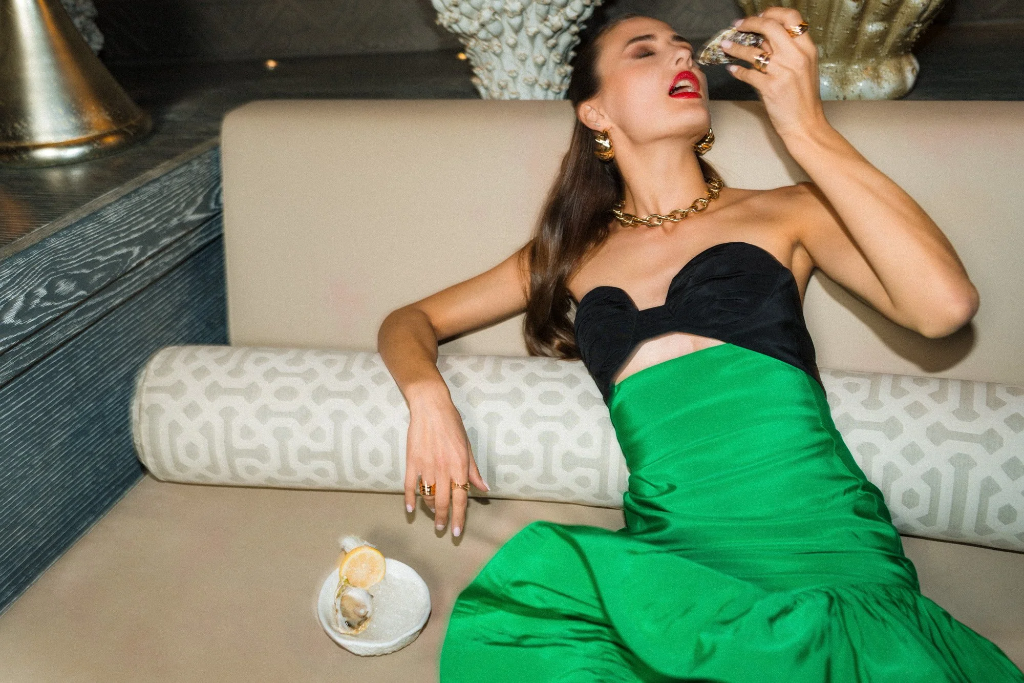 A woman in an elegant strapless black and green dress with gold jewelry is reclining on a sofa, holding a glass of champagne near her mouth, with a drink on a small table beside her.
