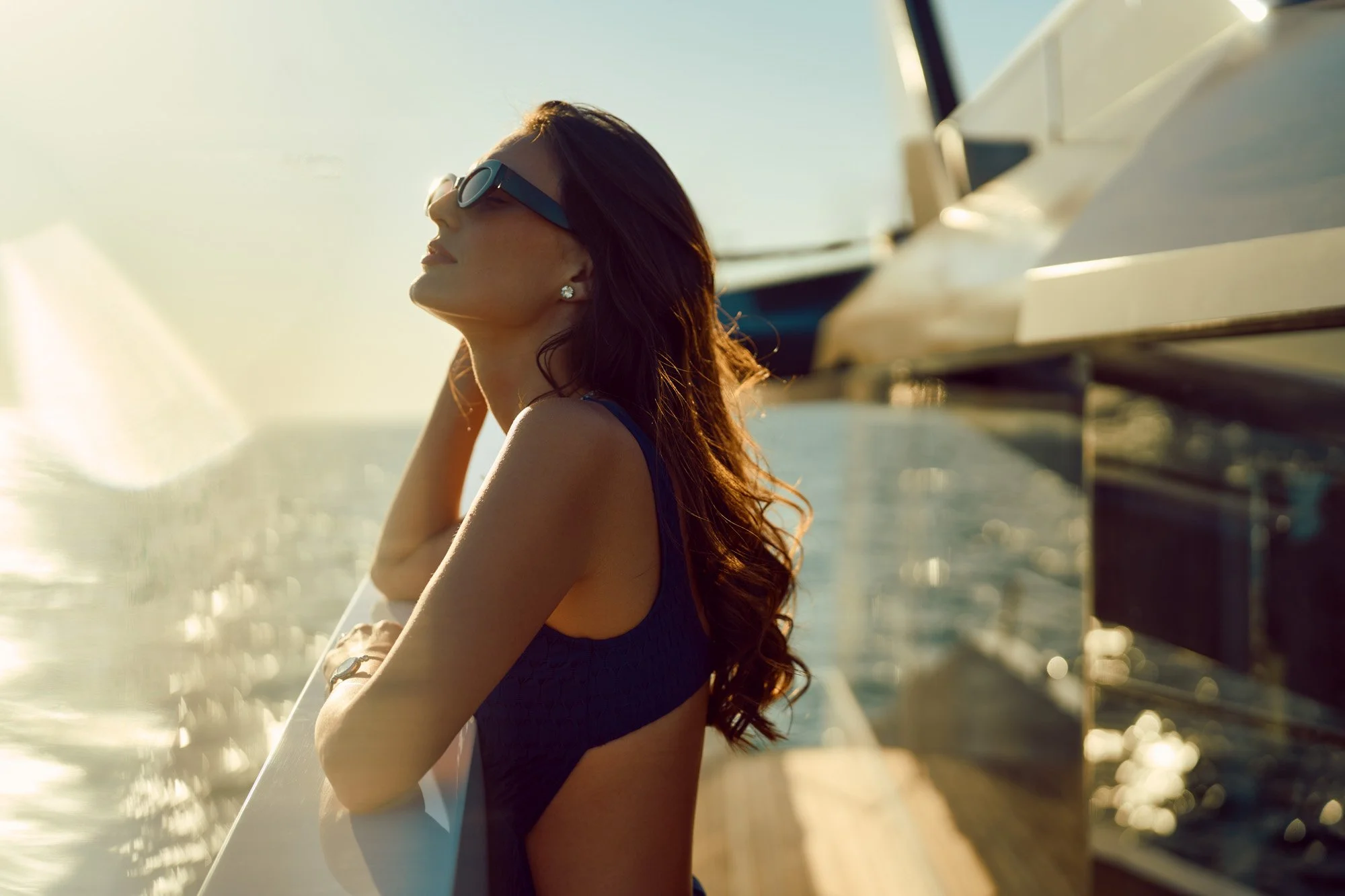 A woman with long dark hair, wearing sunglasses and a blue swimsuit, relaxes on a yacht at sunset.