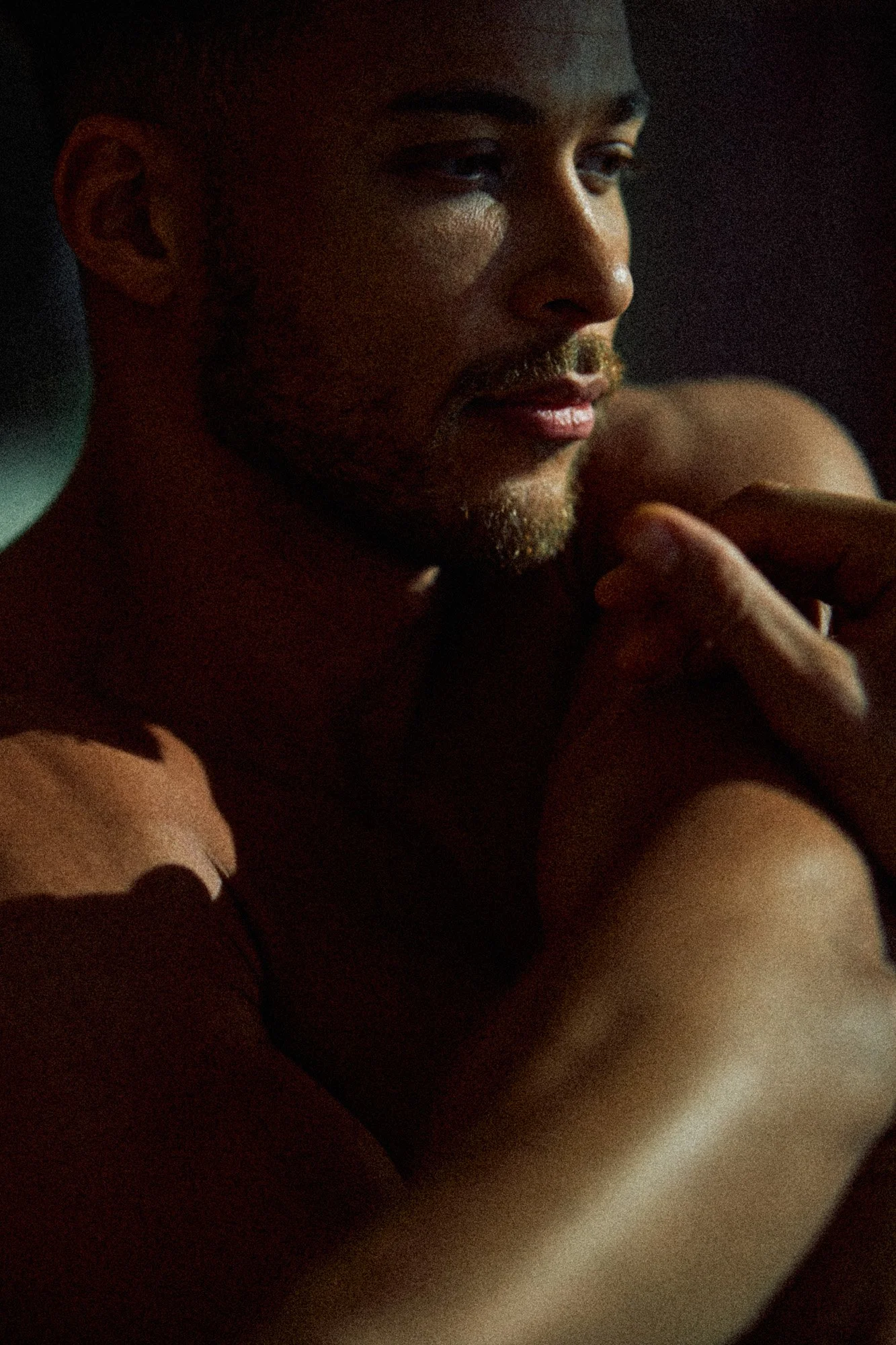 A close-up of a shirtless man with a beard and short hair, resting his chin on his hand, in a dimly lit setting.