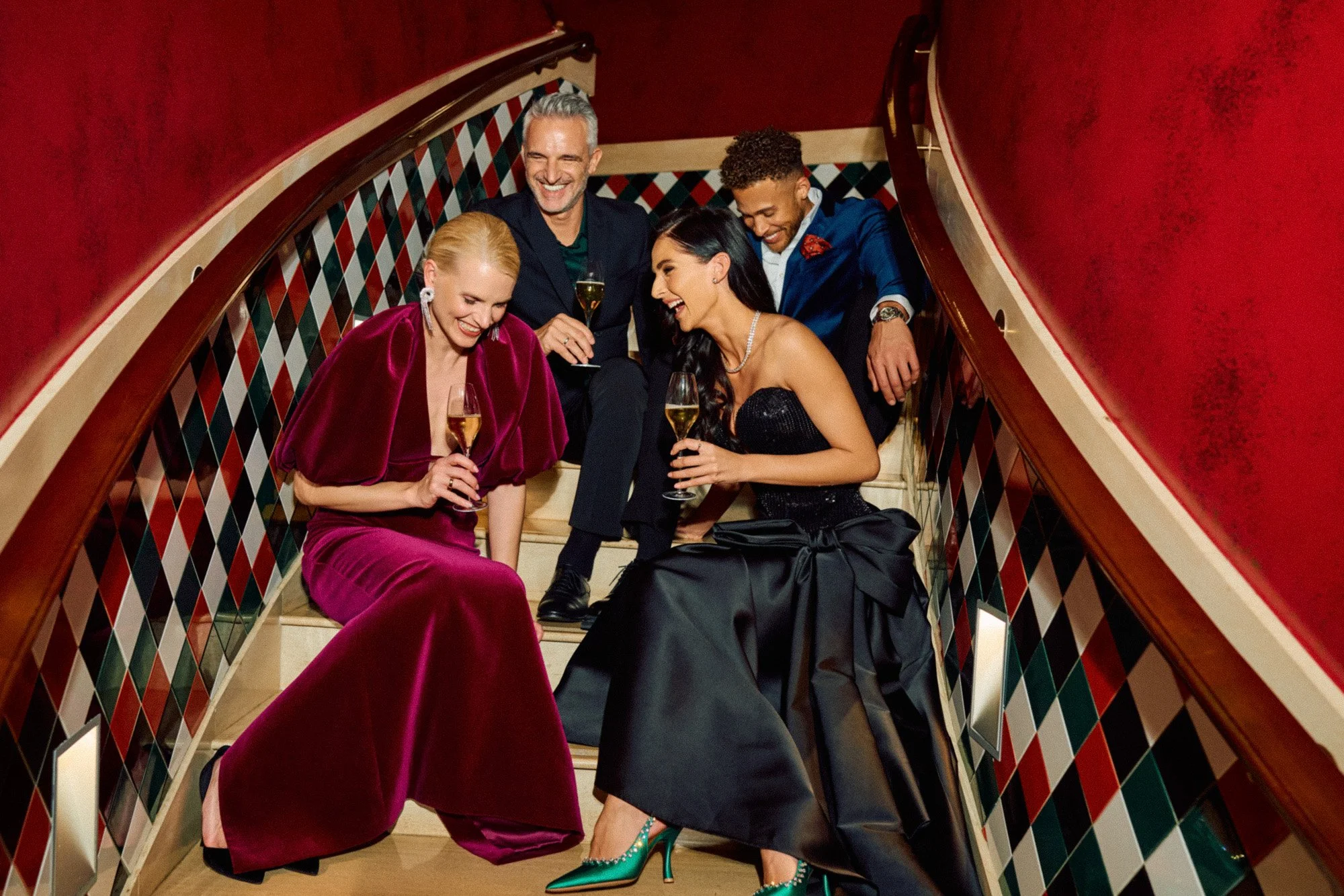 A group of five well-dressed people laughing and talking while sitting on a staircase with colorful, patterned tiles, holding glasses of champagne.