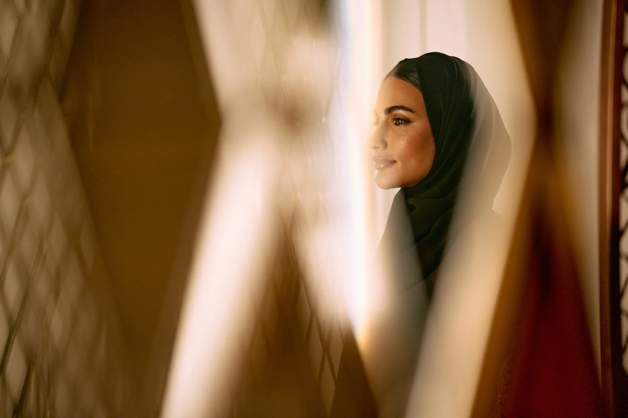 A woman wearing a black hijab looking to the side, seen through a decorative mirror with gold frames.