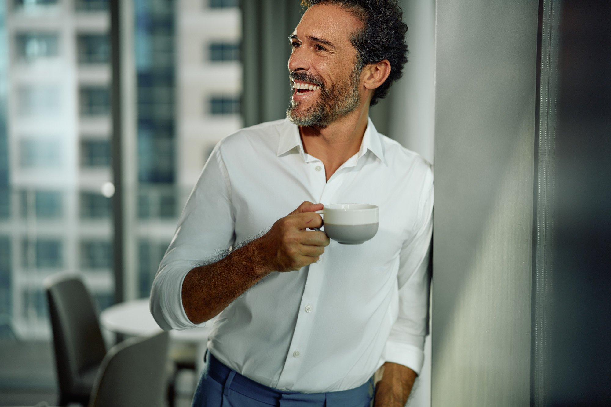 A middle-aged man with dark, curly hair and a beard, smiling while holding a white cup, standing near a window with a cityscape background.