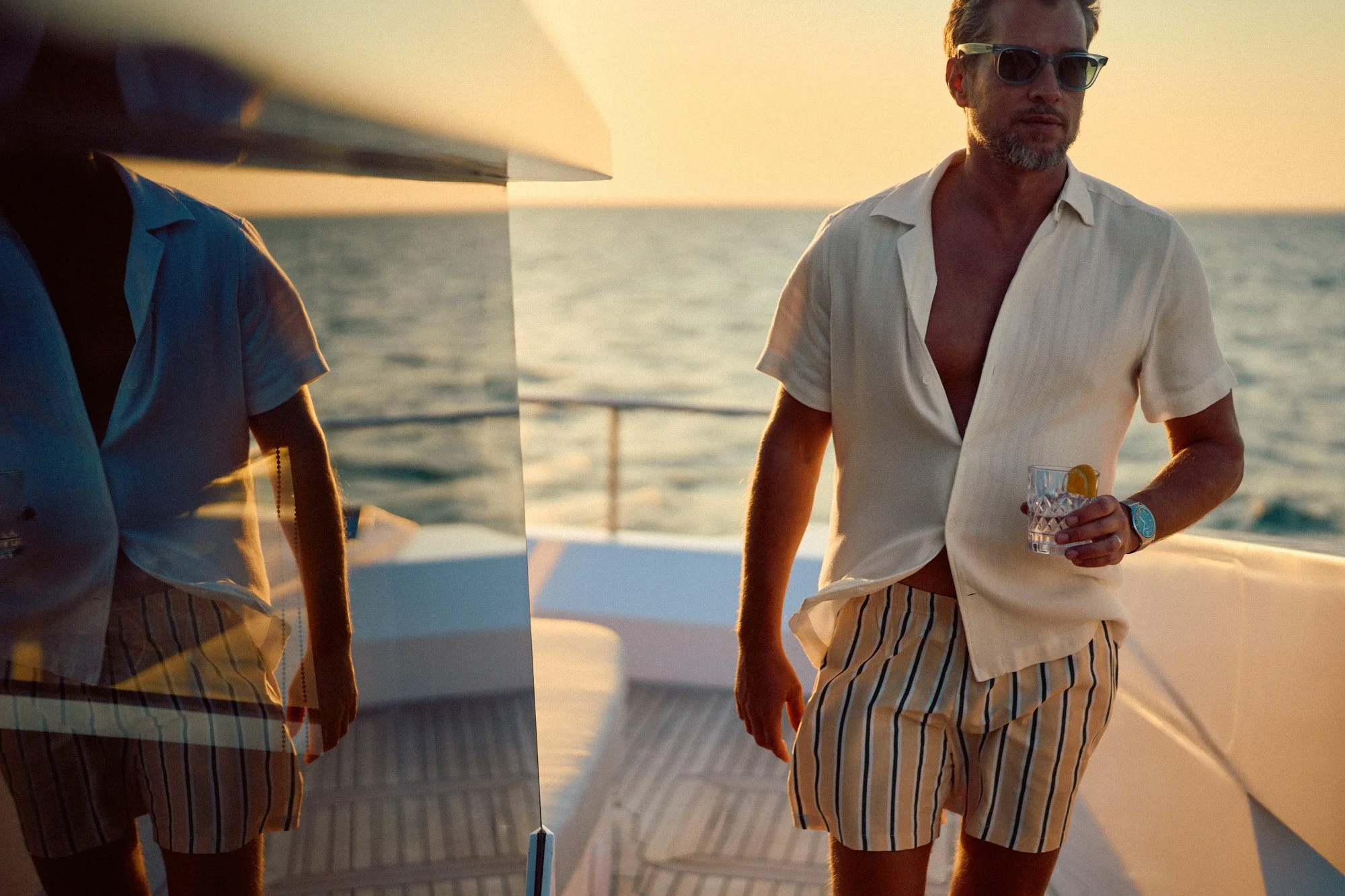 A man on a yacht at sunset, wearing sunglasses, an open short-sleeve button-up shirt, and striped shorts, holding a drink with lemon.