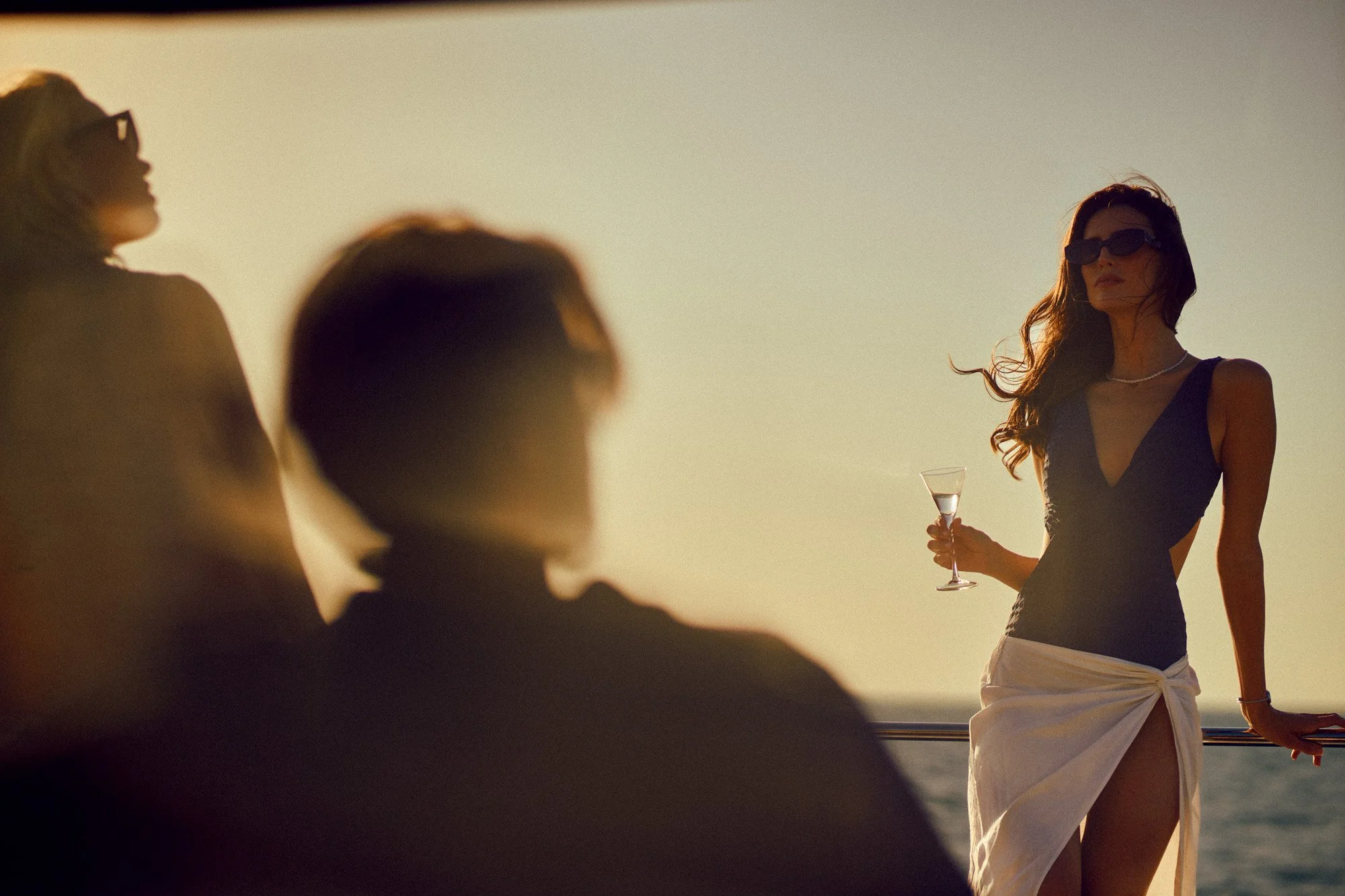 Woman in black dress and sunglasses holding a martini glass on a boat at sunset, with two blurred figures in the foreground.