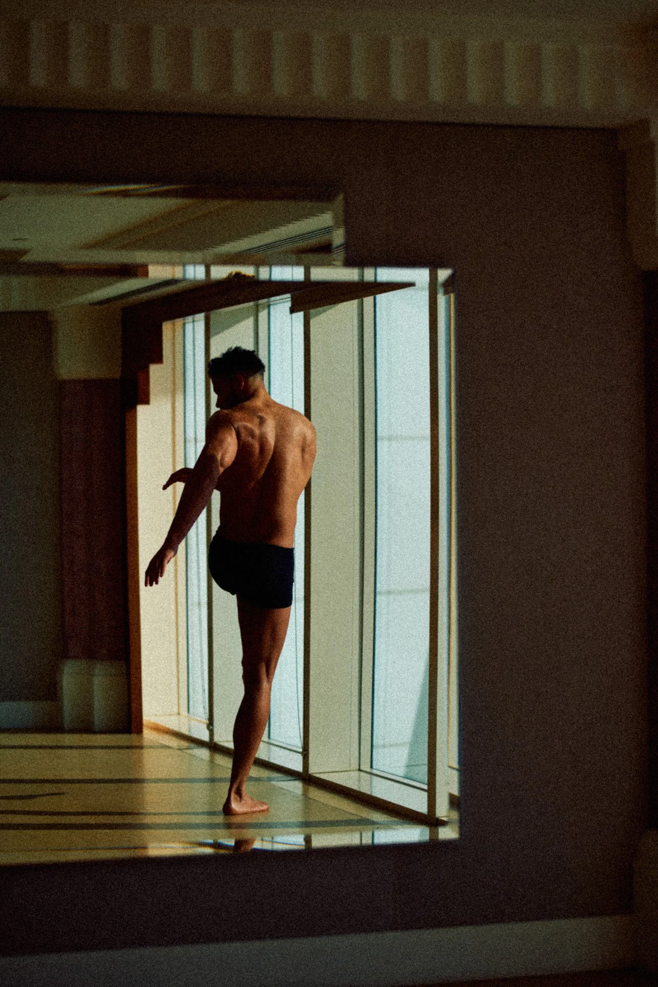 A man standing in front of a large window, wearing black shorts, stretching or adjusting his arm.